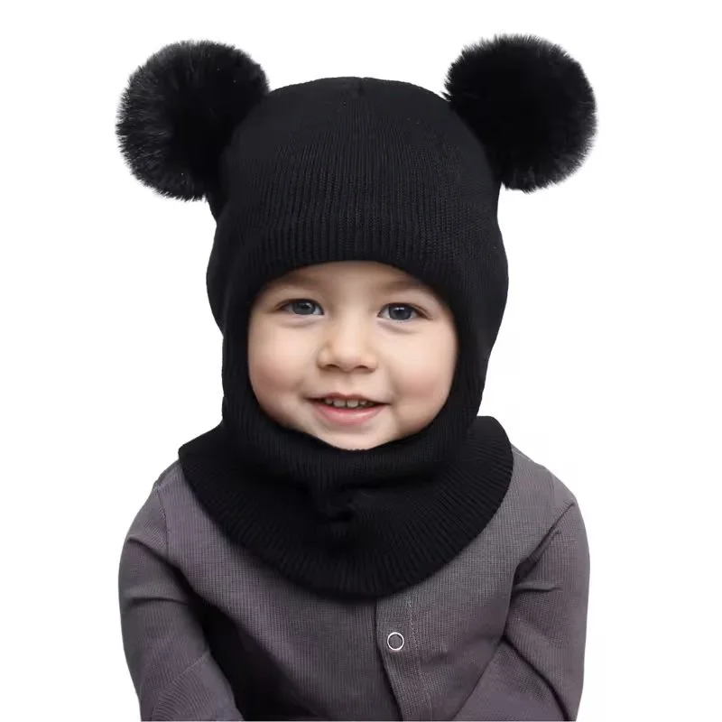 A young child wearing a black knit hat with two fluffy pom-poms and a matching neck warmer, smiling at the camera.