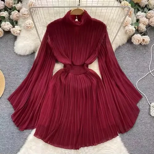 A red pleated midi dress with long sleeves and a high neckline displayed on a hanger, surrounded by flowers and furry rugs.