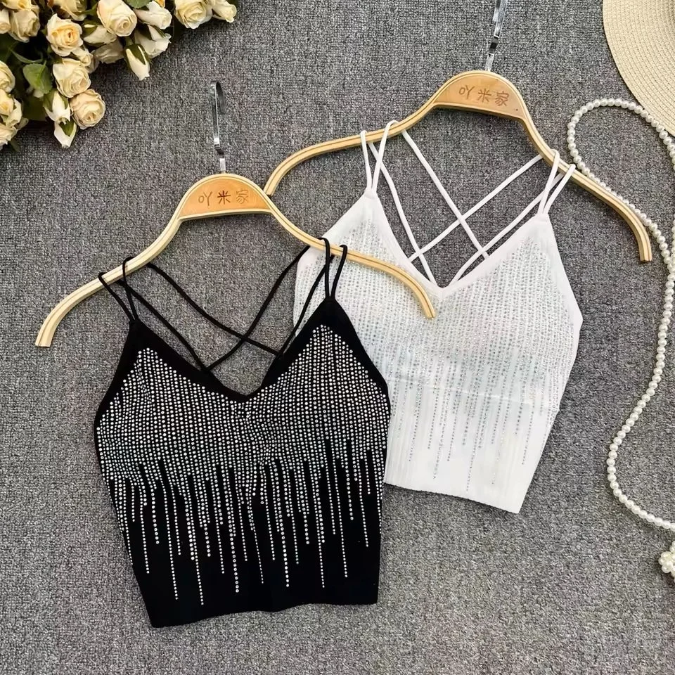 Two stylish cropped tops with thin straps, one black with rhinestone embellishments and the other white with similar rhinestone details, displayed on hangers on a gray surface. Decorative flowers and a pearl necklace are also visible.