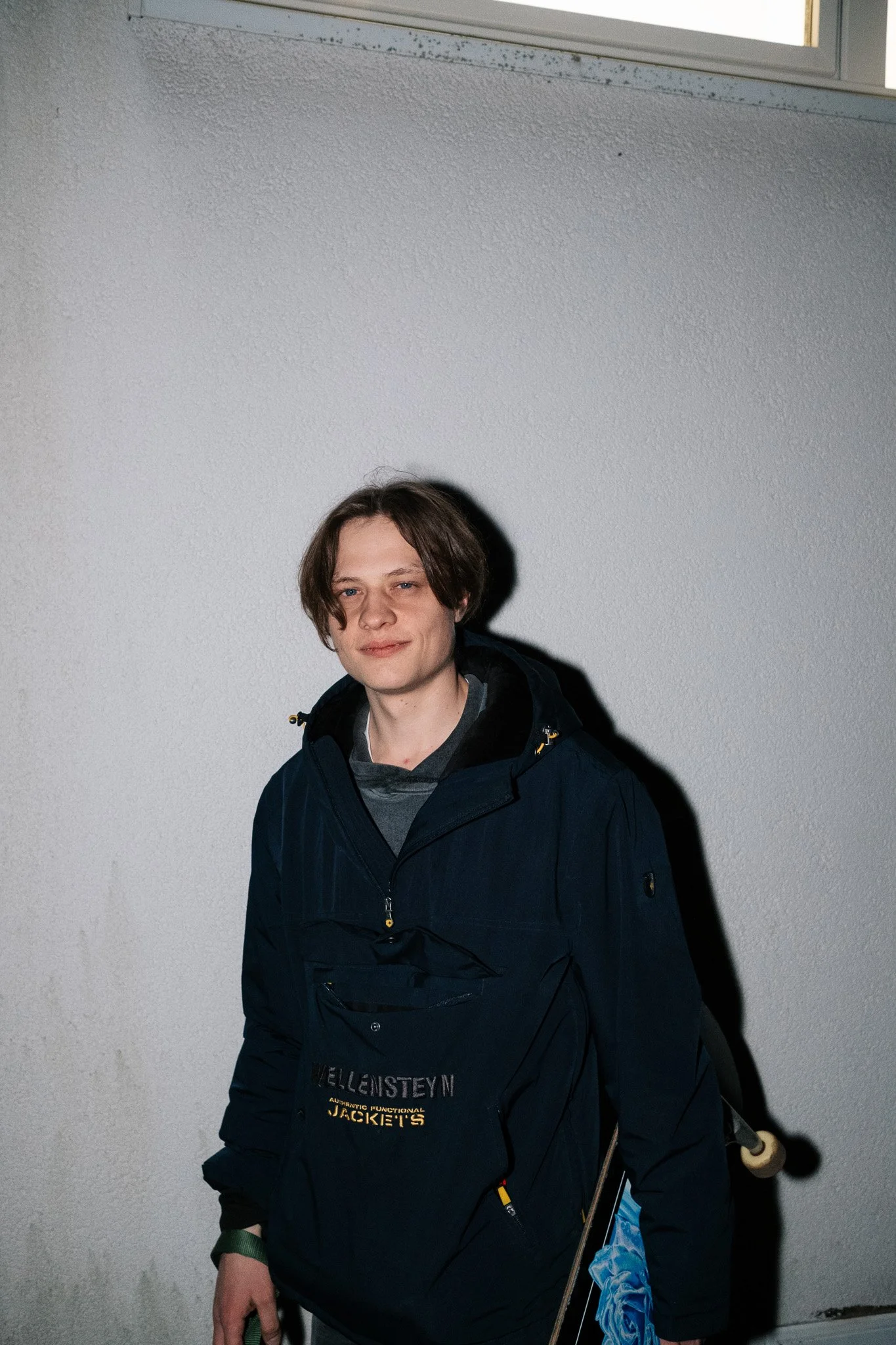 Junge Person mit braunen Haaren trägt eine dunkle Jacke und hält eine Bambusflöte, steht vor einer weißen Wand.