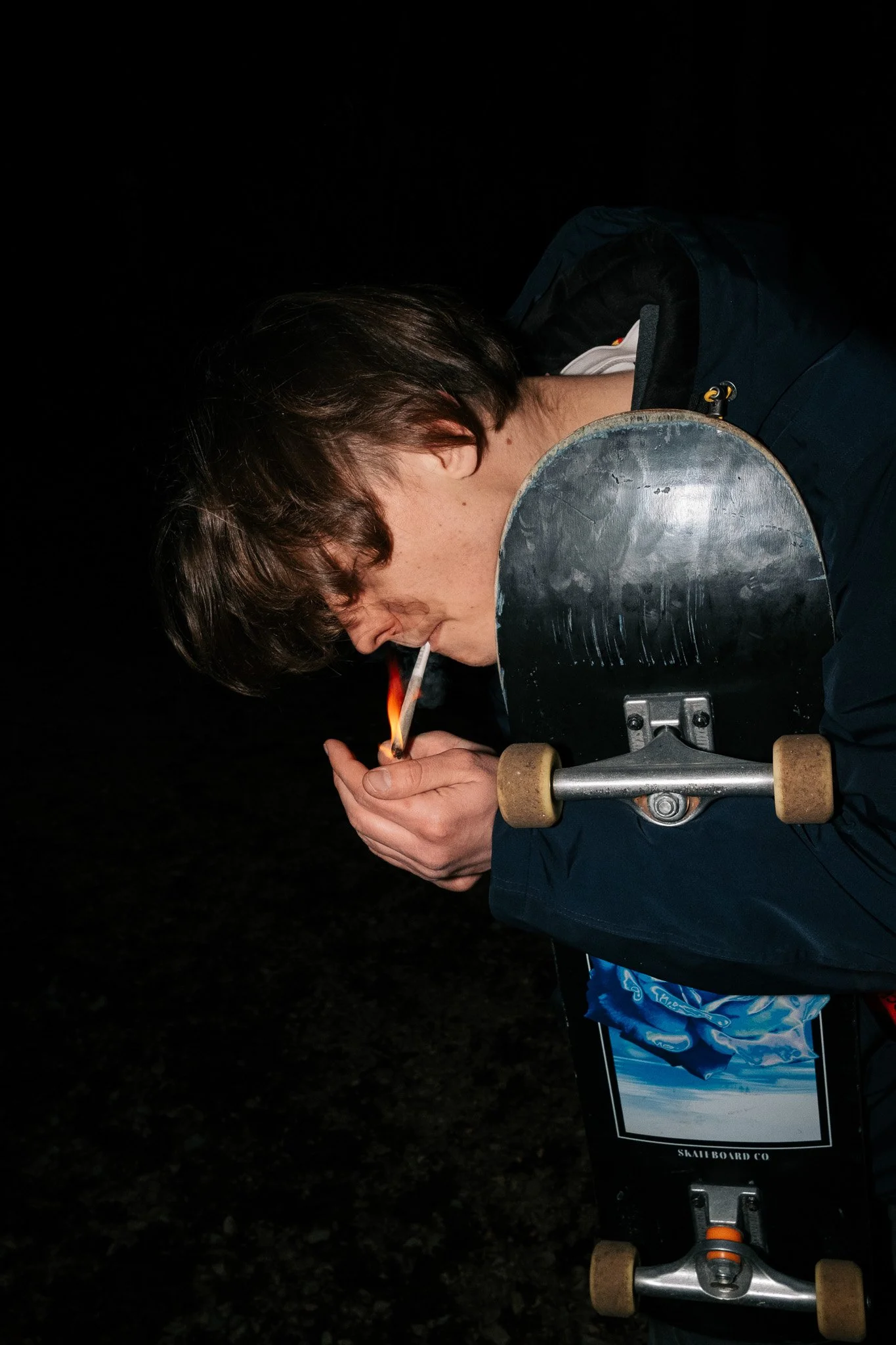 Eine Person zündet eine Zigarette an, während sie an einem Skateboard lehnt.