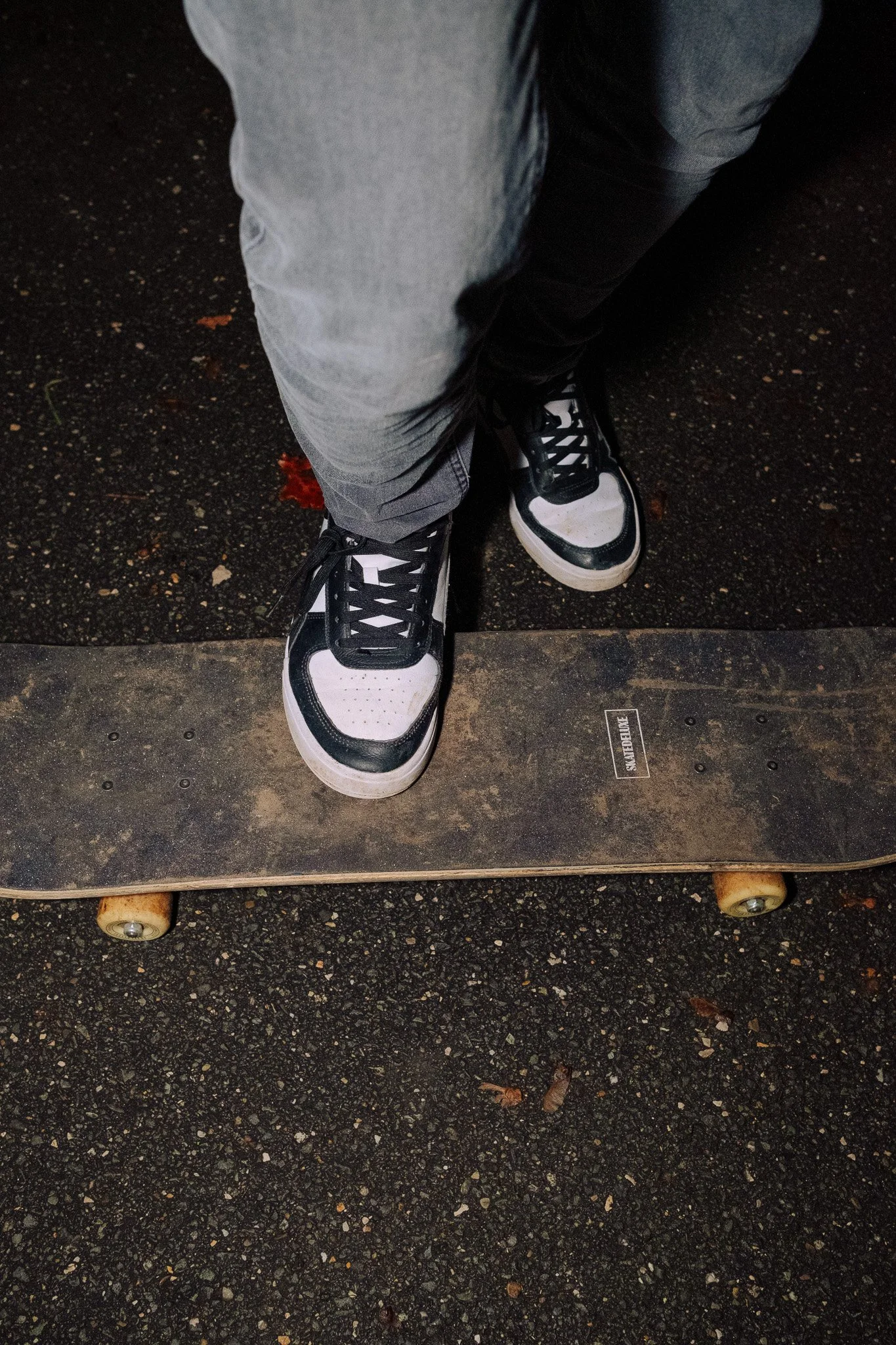 Nahaufnahme von schwarzen und weißen Turnschuhen auf einem Skateboard, getragen von einer Person, die graue Hosen trägt, auf einer asphaltierten Oberfläche.