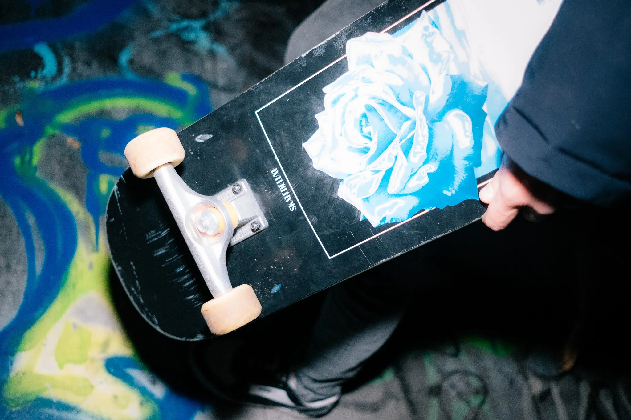Nahaufnahme eines Skateboards mit blauem und weißen Print, das von einer Person mit schwarzer Jacke gehalten wird, die auf einem graffitiübermalten Boden steht.