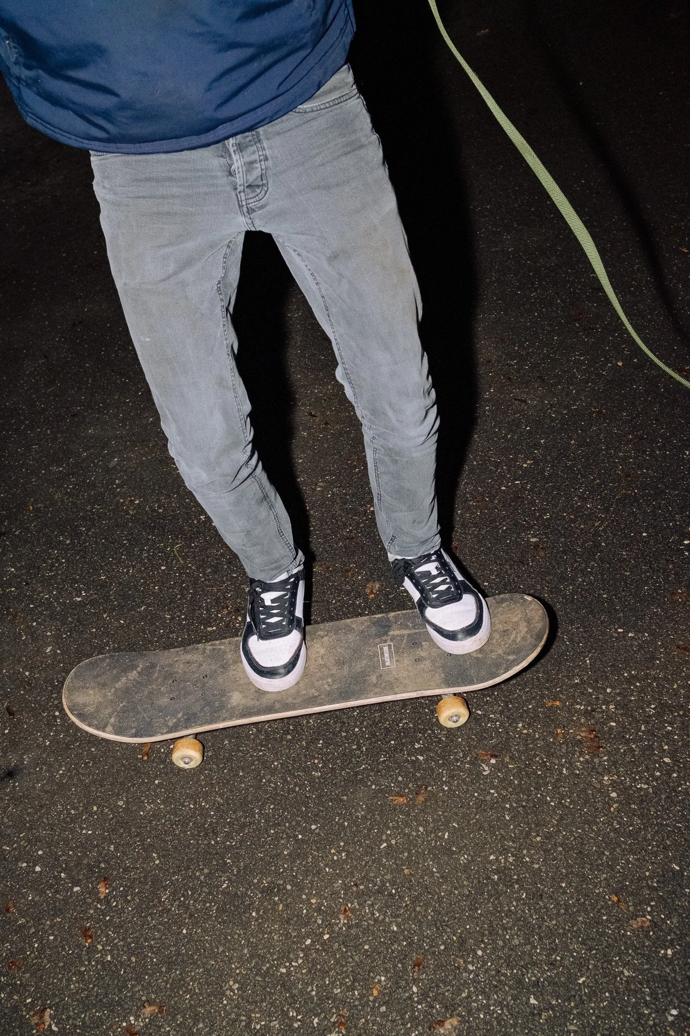 Person auf einem Skateboard, getragen mit schwarzen und weißen Turnschuhen, auf asphaltartigem Boden bei Nacht.