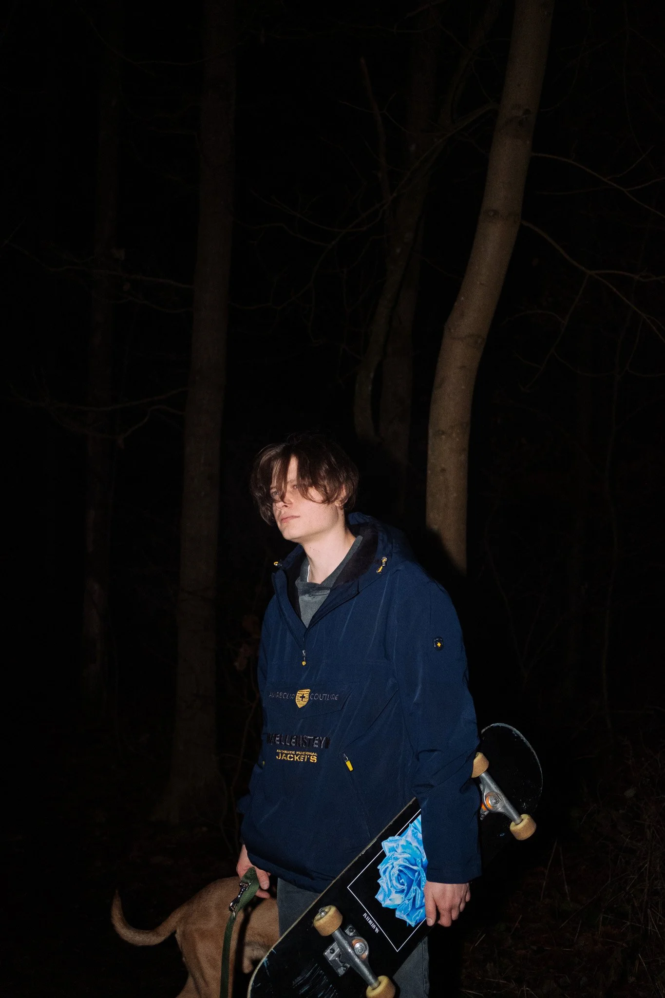 Junge mit dunklem Haar, blauer Jacke, hält Skateboard und Hund, steht im dunklen Wald bei Nacht.