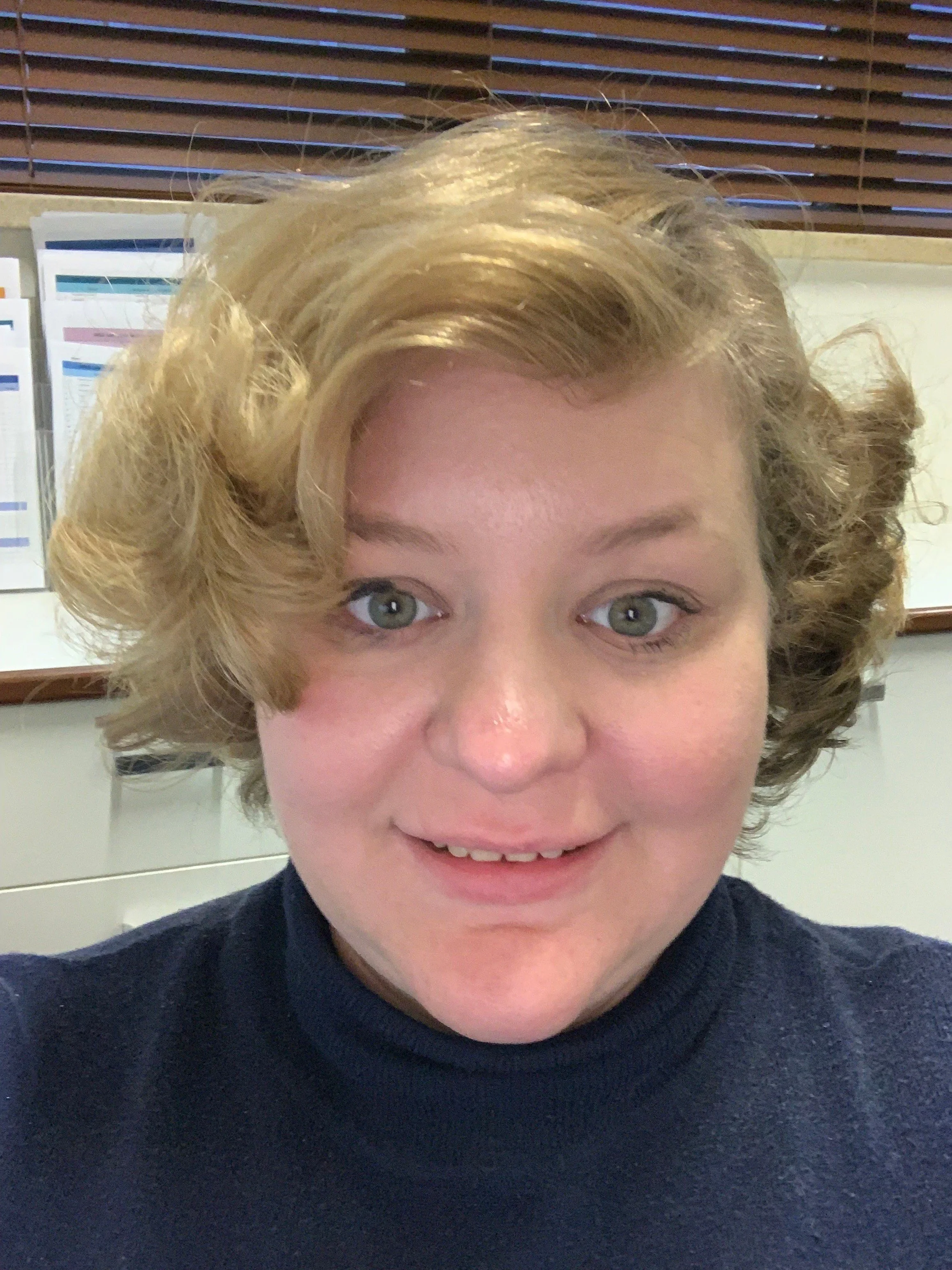 A woman with short, curly blonde hair and gray eyes smiling at the camera, wearing a black turtleneck, sitting in an office with window blinds and documents in the background.
