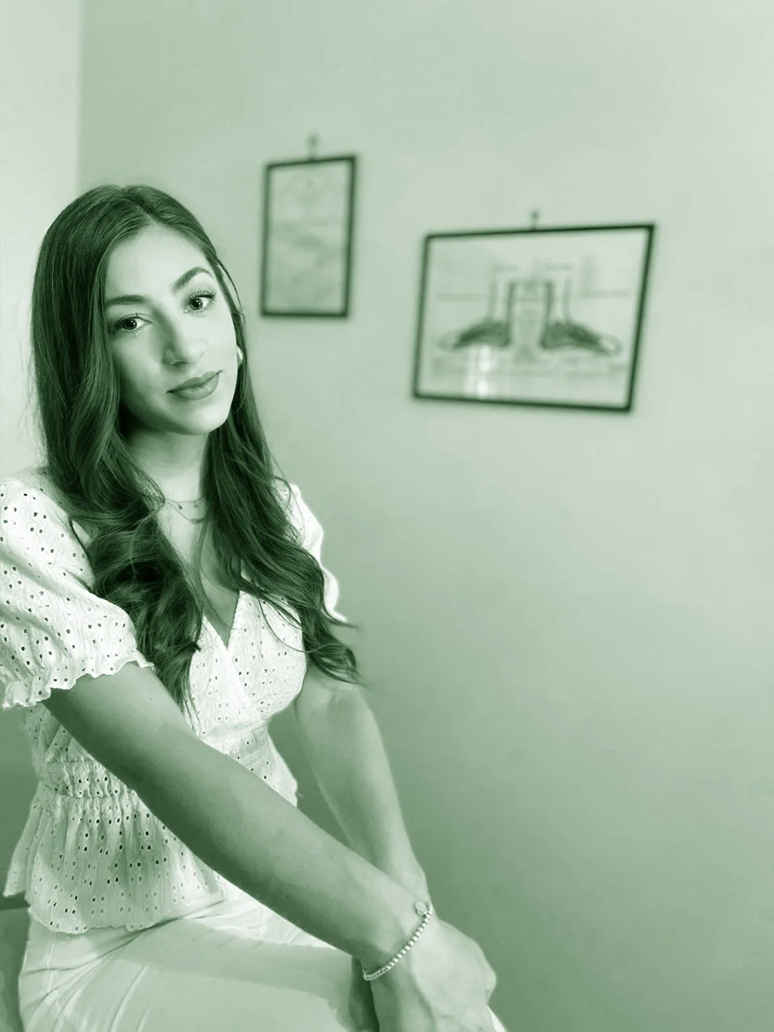 Una donna seduta su una sedia, con capelli lunghi e ricci, abbigliamento chiaro, con un braccialetto, sfondo con due quadri appesi al muro.