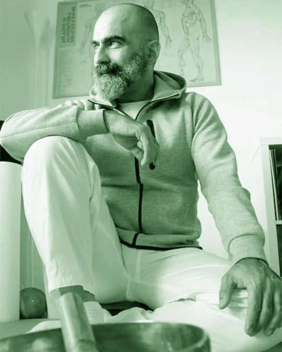 Uomo con barba e testa rasata, indossa una felpa grigia e pantaloni bianchi, seduto in un ambiente con poster sullo sfondo.