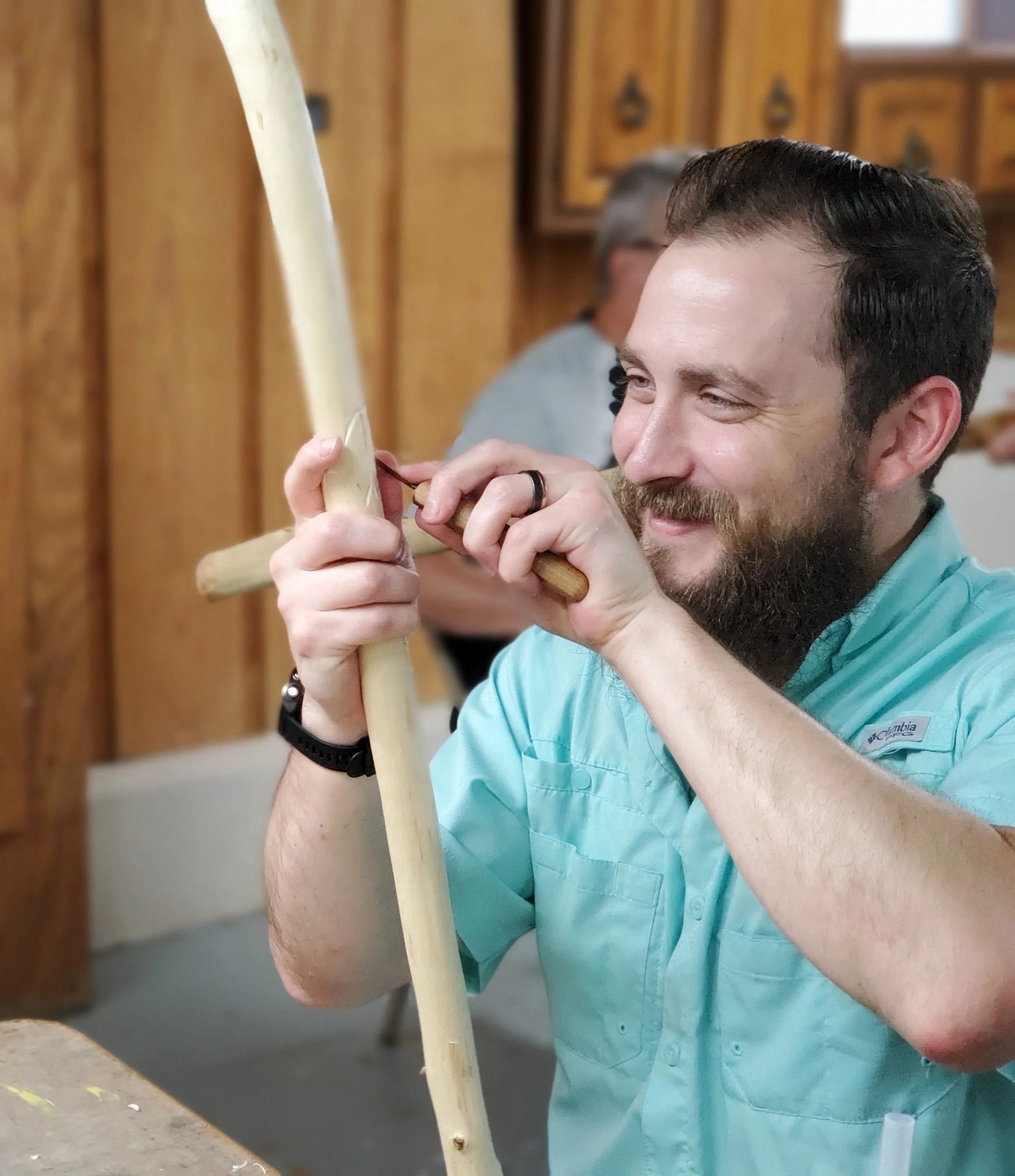 Person with a beard and mustache, wearing a light blue collared shirt, smiling while carving or shaping a piece of light-colored wood with a hand tool inside a room with wooden cabinets in the background.