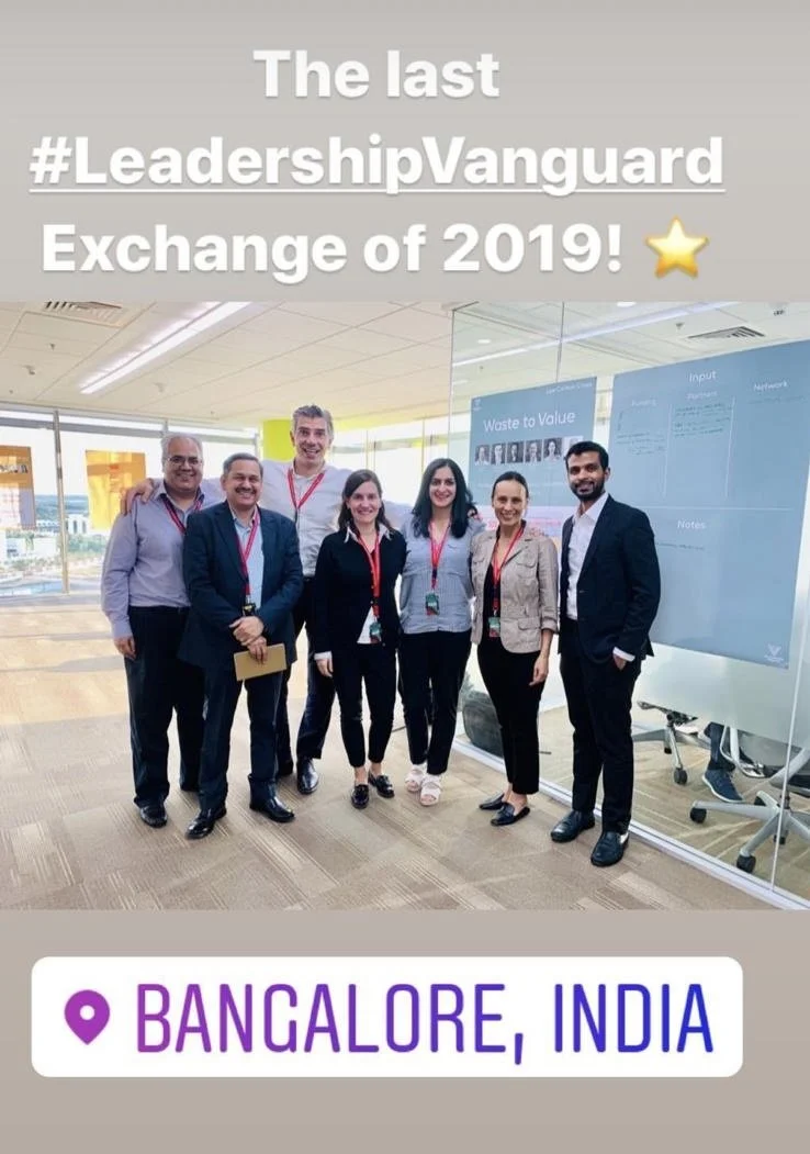 Group of seven professionals posing in a conference room at Bangalore, India, during the Leadership Vanguard Exchange of 2019, with presentation boards in the background and all wearing conference badges.