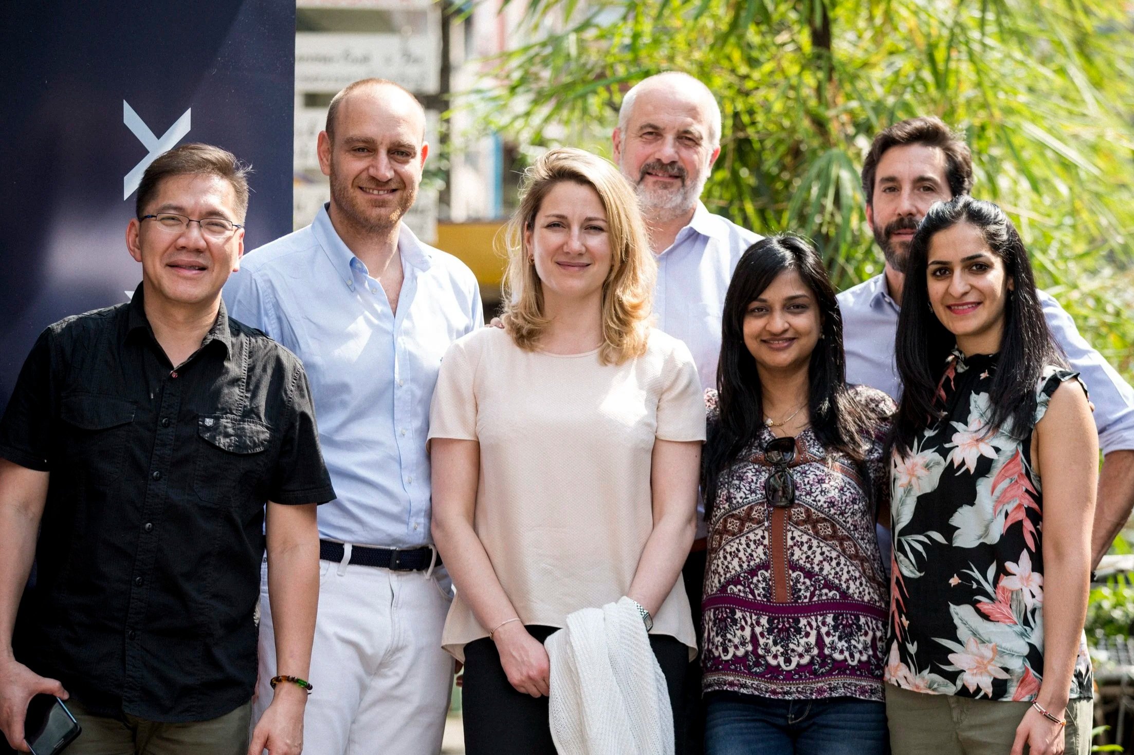 Group of eight diverse people standing outdoors, smiling for the camera, with greenery in the background.