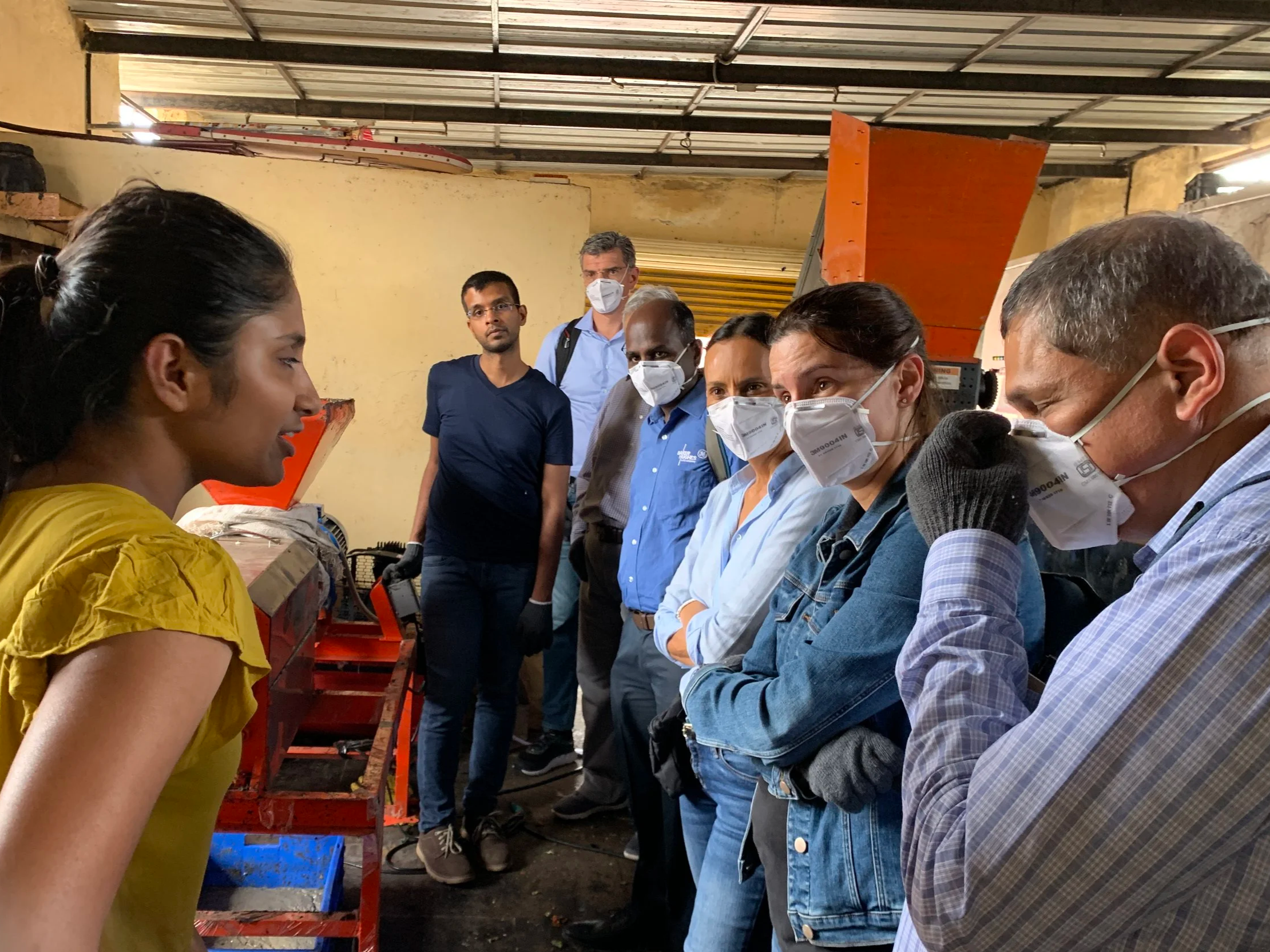 A group of people, mostly wearing masks, observing a young woman demonstrating or explaining something in an industrial or workshop setting.