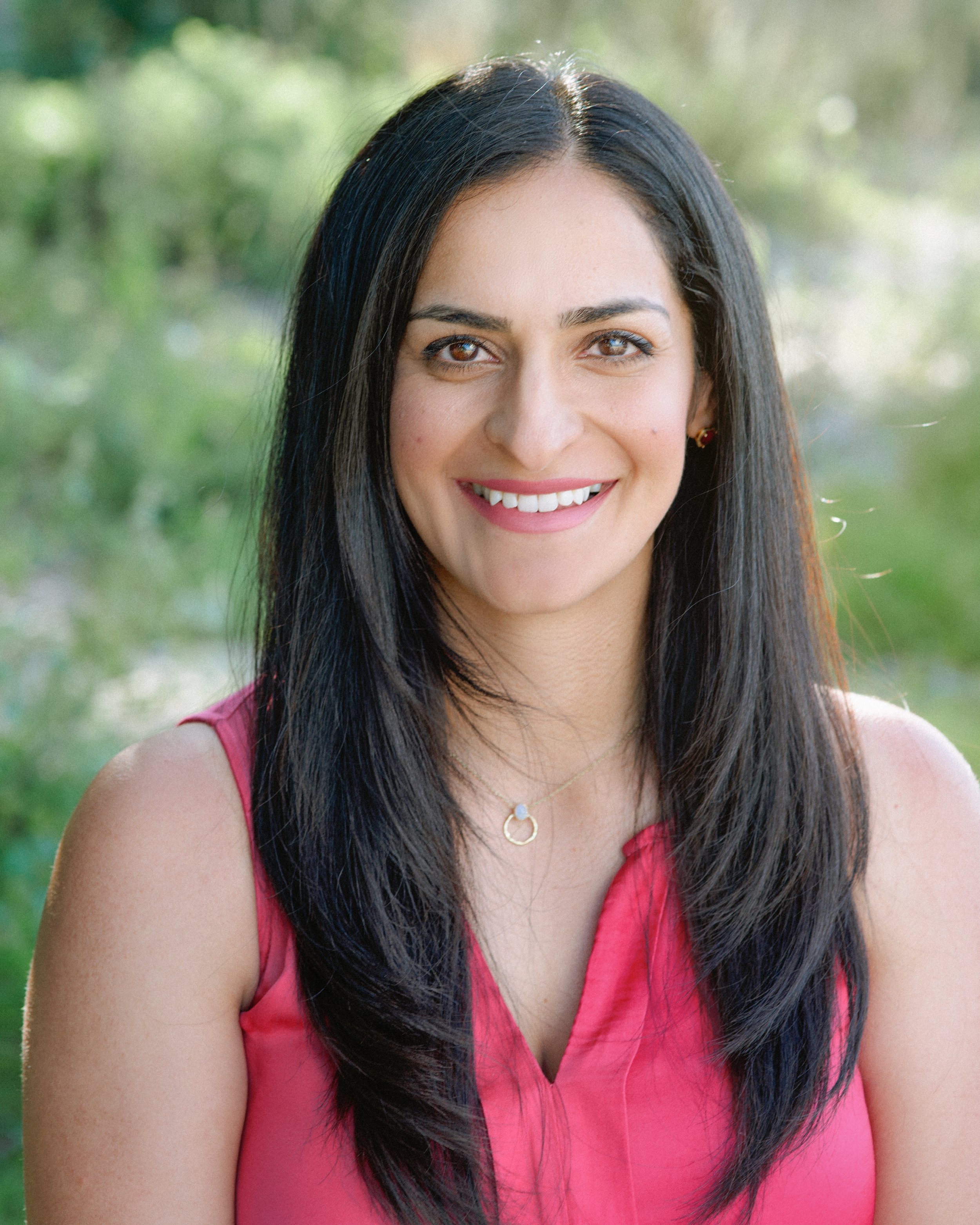 A woman with long black hair, wearing a pink sleeveless top and jewelry, smiling outdoors with a blurred green background.