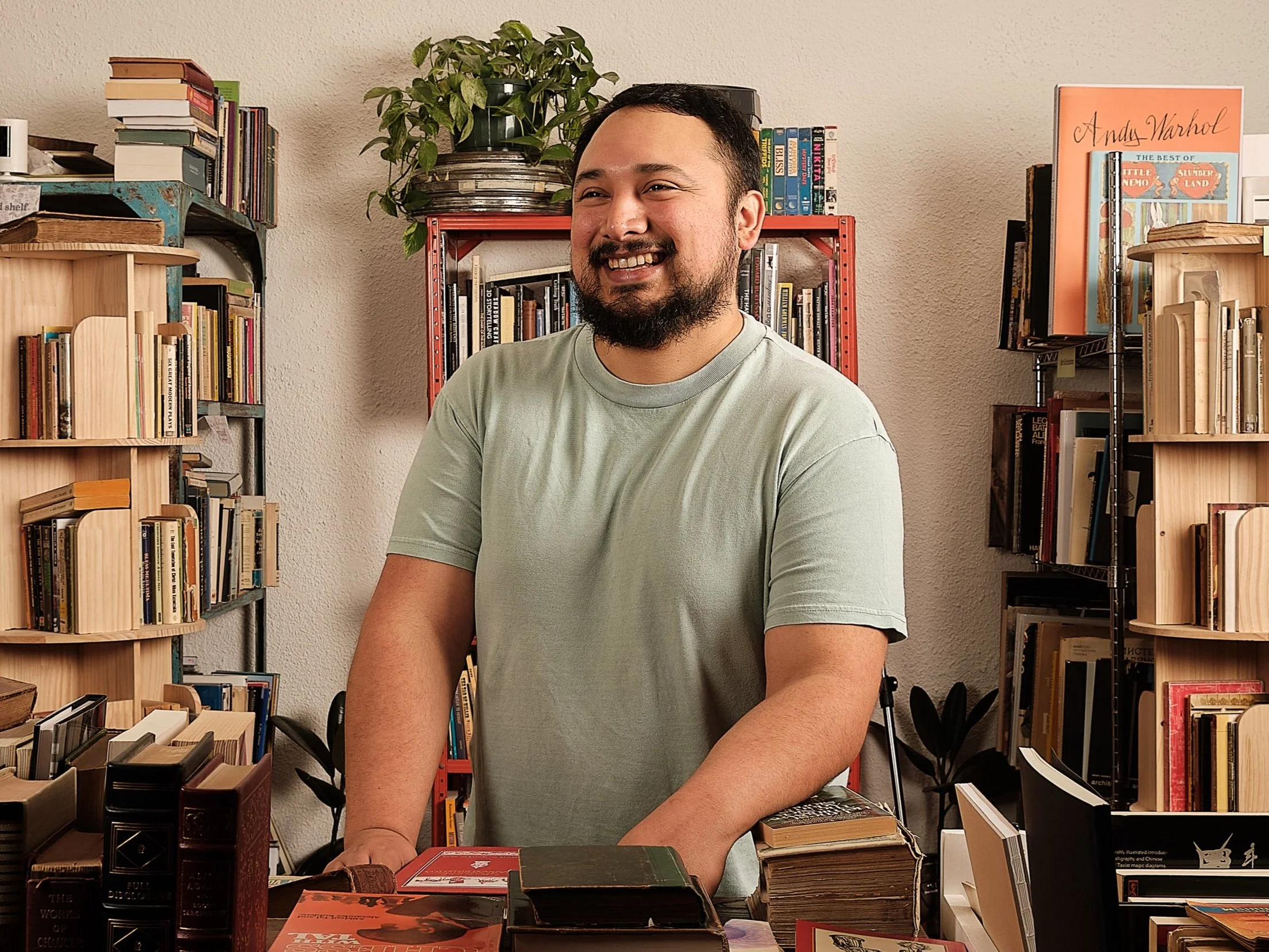 A smiling man with a beard and short dark hair, wearing a light green t-shirt, standing behind a table filled with books, in a room with bookshelves and a potted plant in the background.
