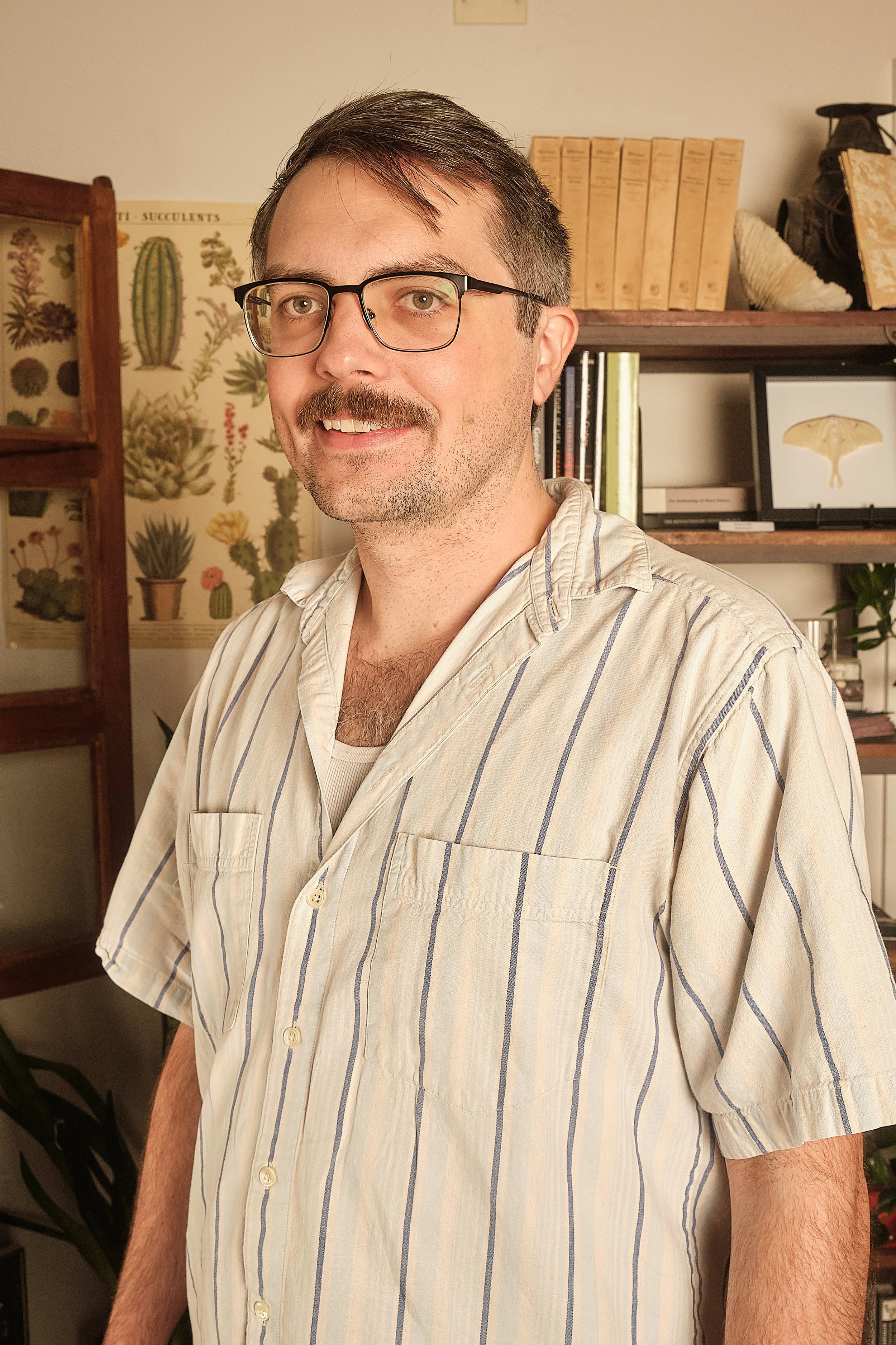 A man with glasses and a mustache wearing a striped shirt standing in a room with books and plant decorations.