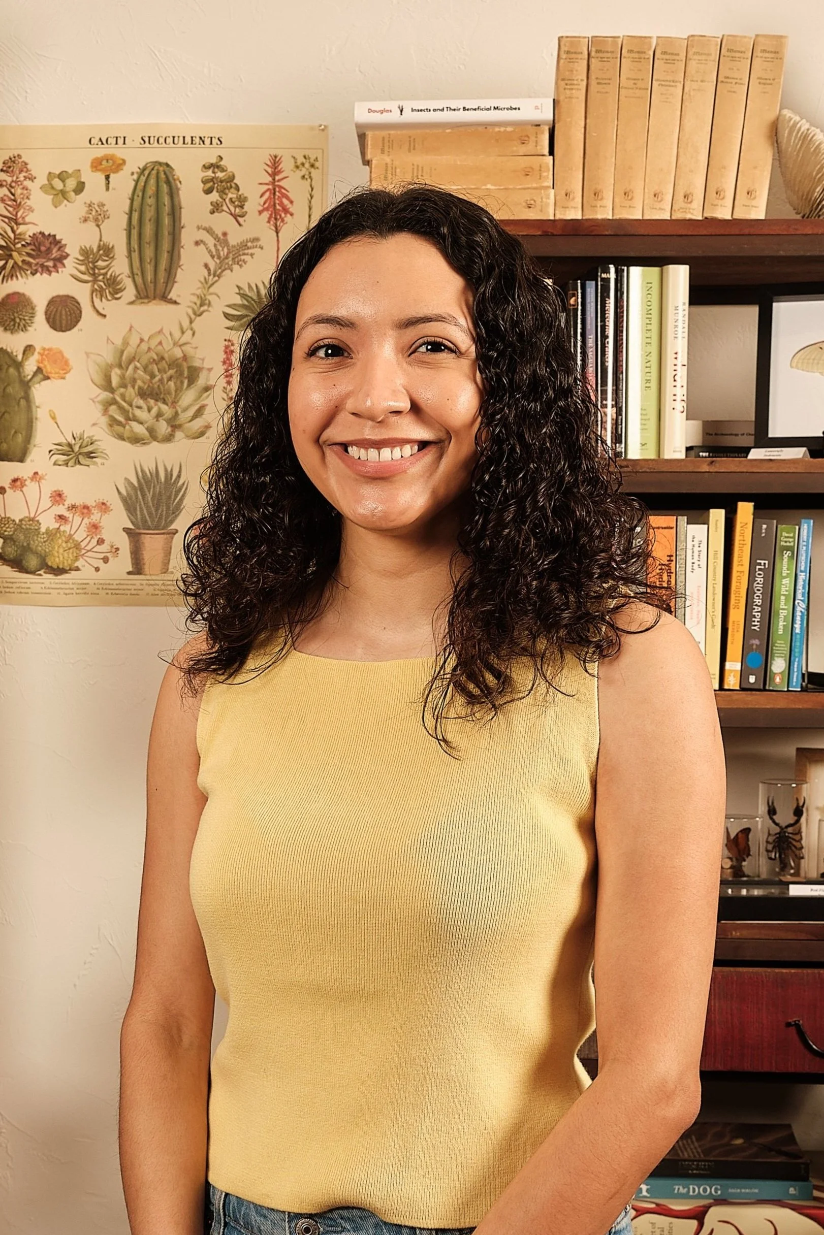 A smiling woman with curly dark hair wearing a yellow sleeveless top standing in front of a bookshelf and a botanical poster