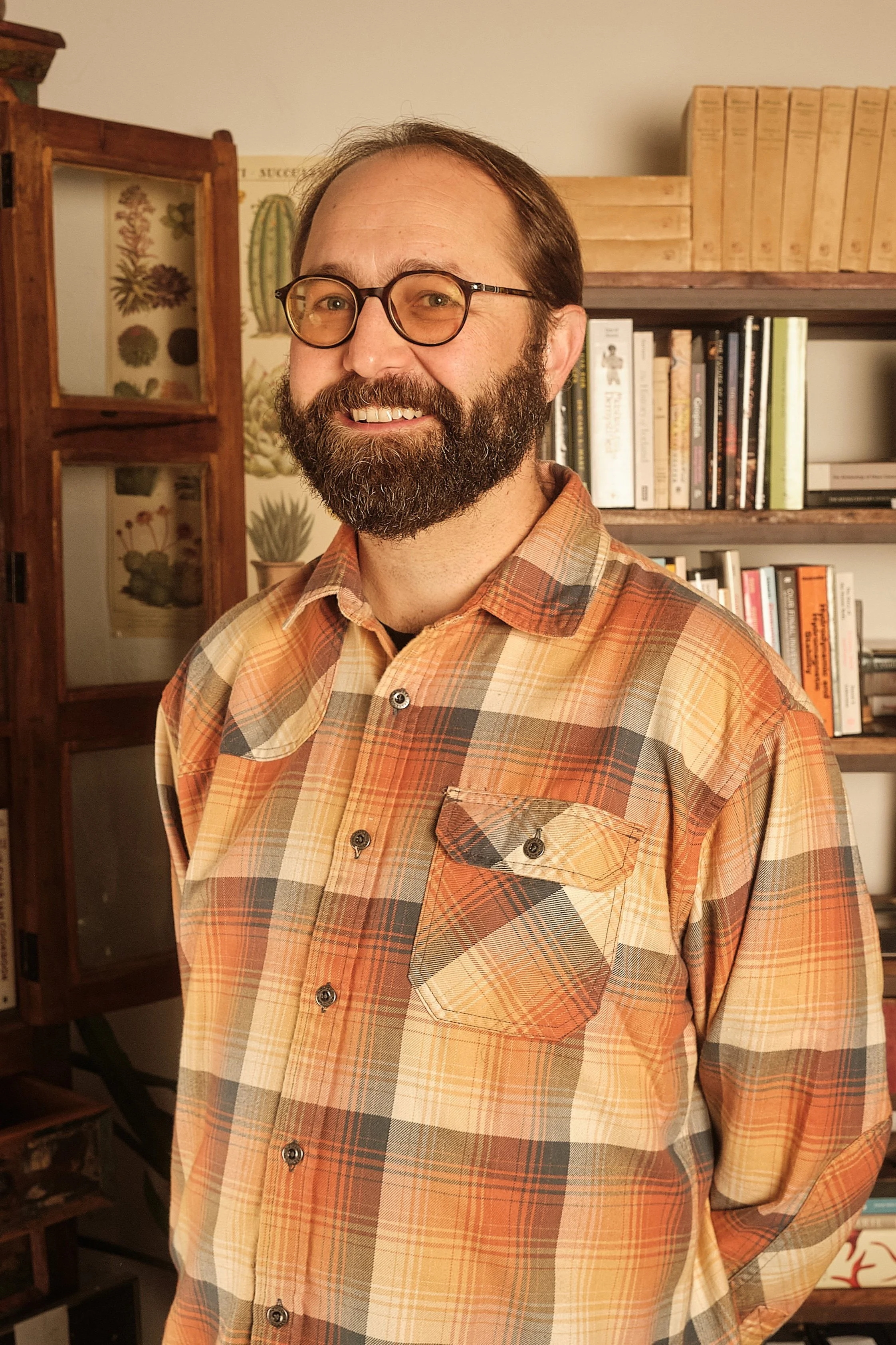 A smiling man with glasses and a beard wearing a plaid shirt standing in a room with bookshelves and plants.