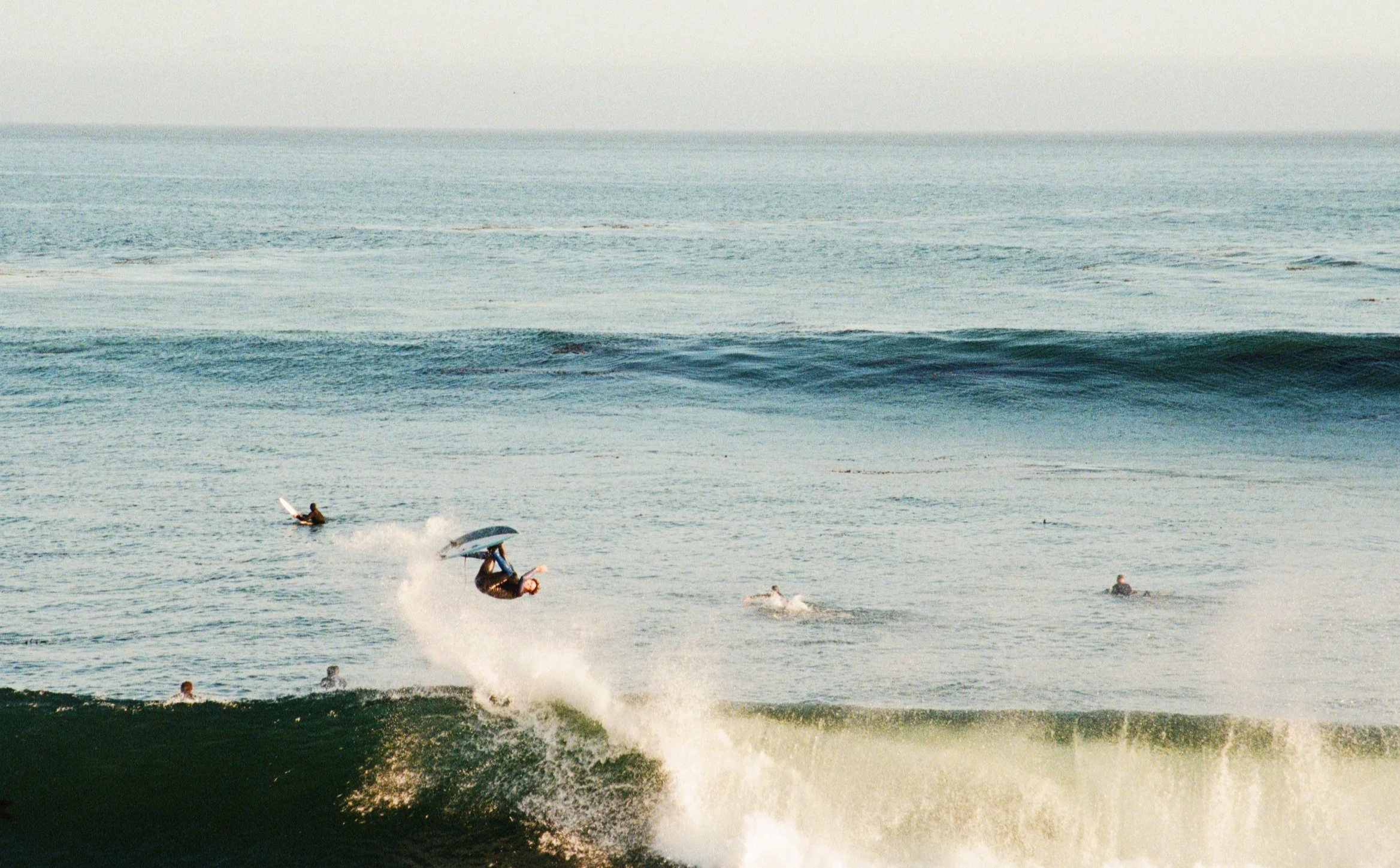Surfer performing a flip above a large wave while others are sitting or swimming in the ocean in the background.