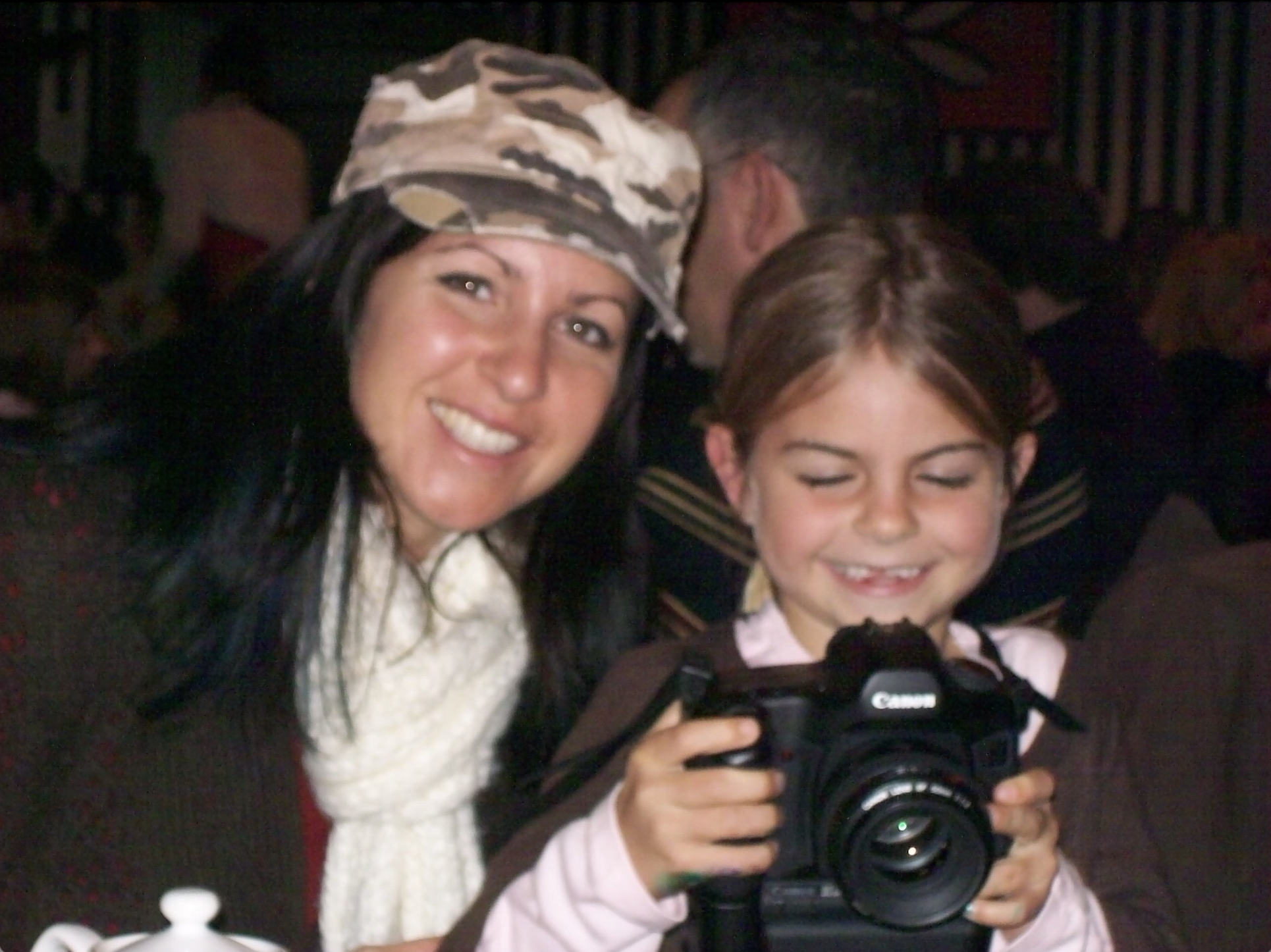 A woman with long black hair wearing a camouflage hat, smiling, next to a young girl with short brown hair holding a camera with both hands, smiling with eyes closed, in a dimly lit room with people in the background.