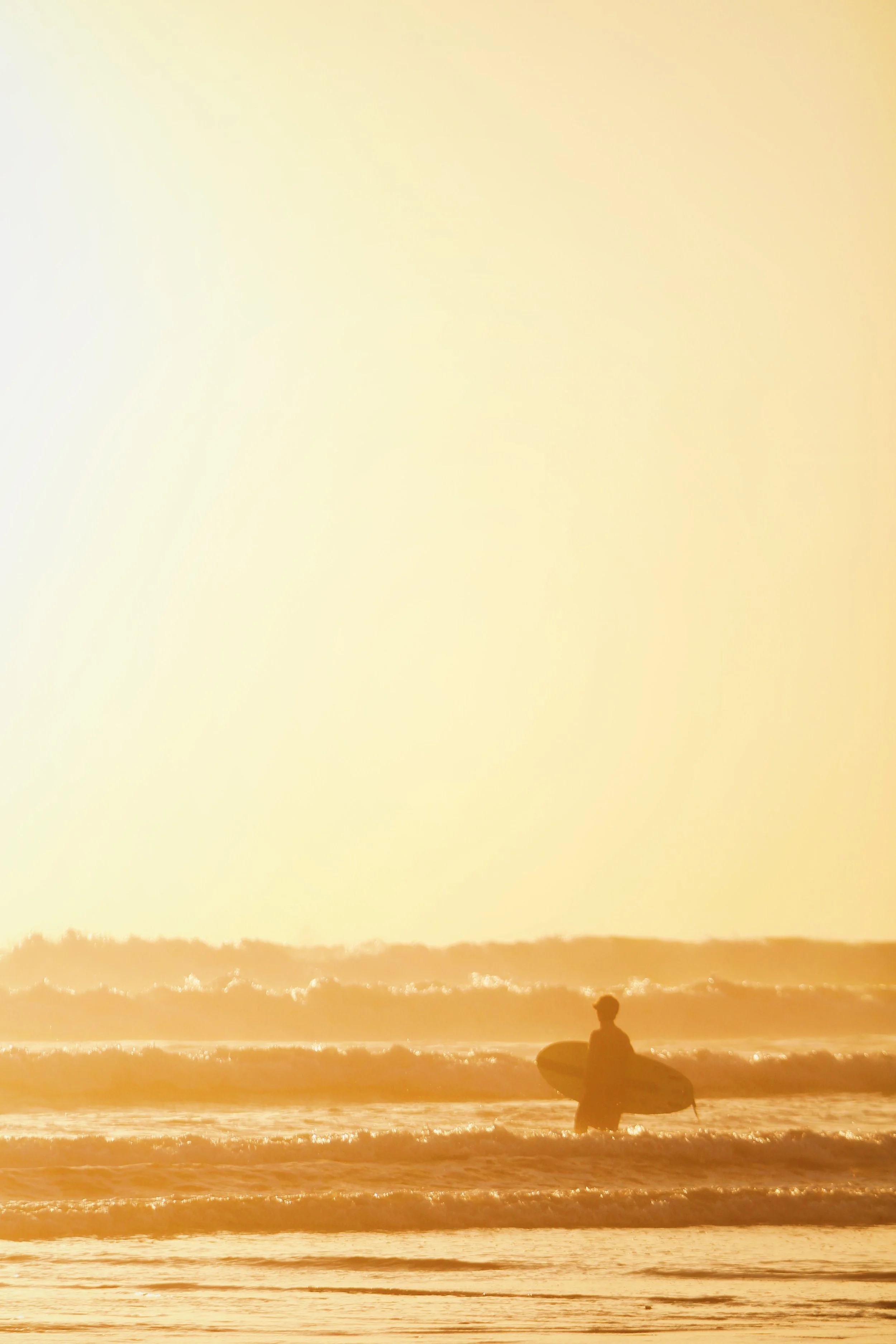 Silhouette of a person with a surfboard standing in the ocean at sunset with waves and a bright yellow sky.