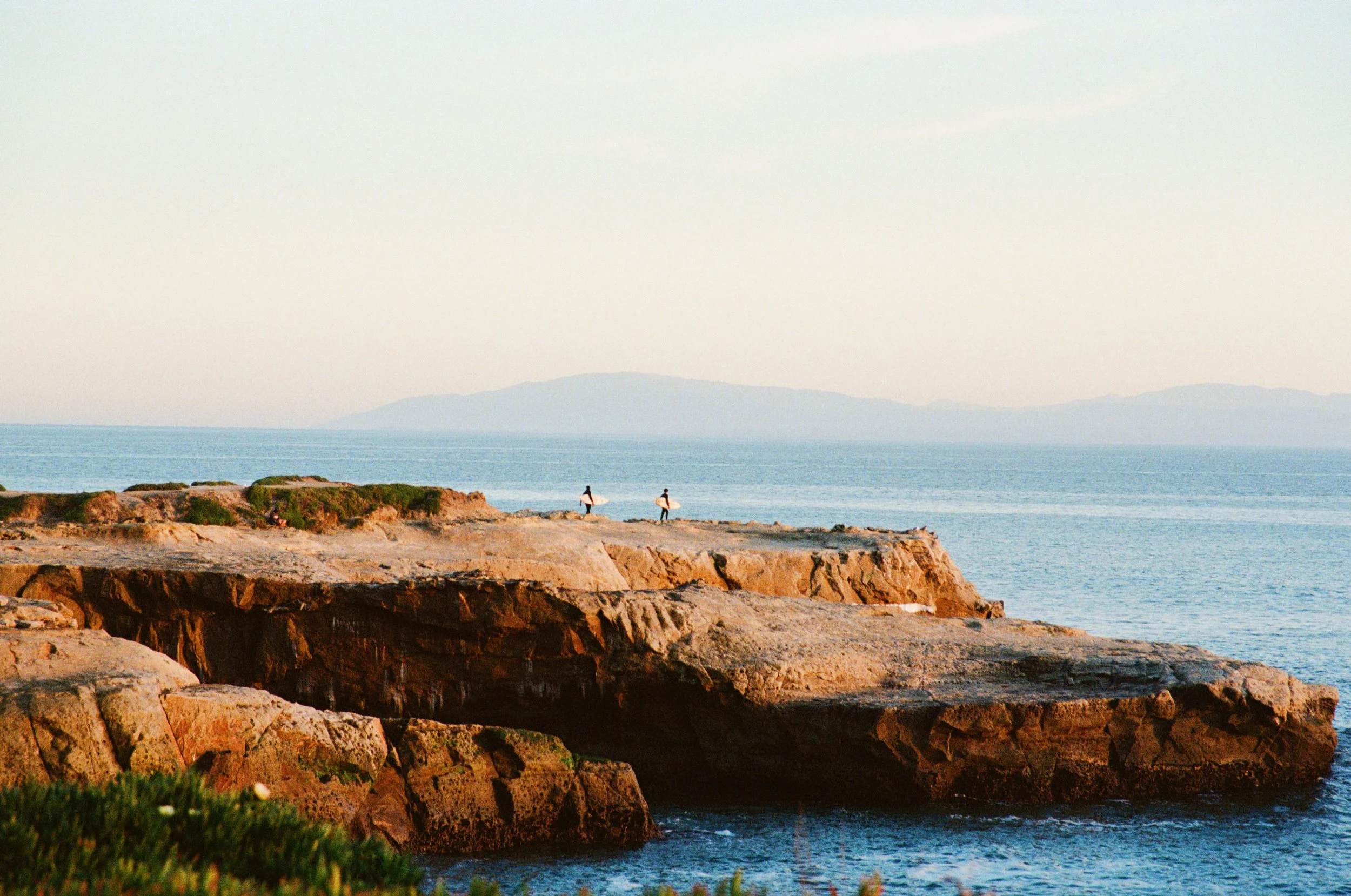 Two surfers walking along rocky coastal cliffs near the ocean with a distant mountain in the background.