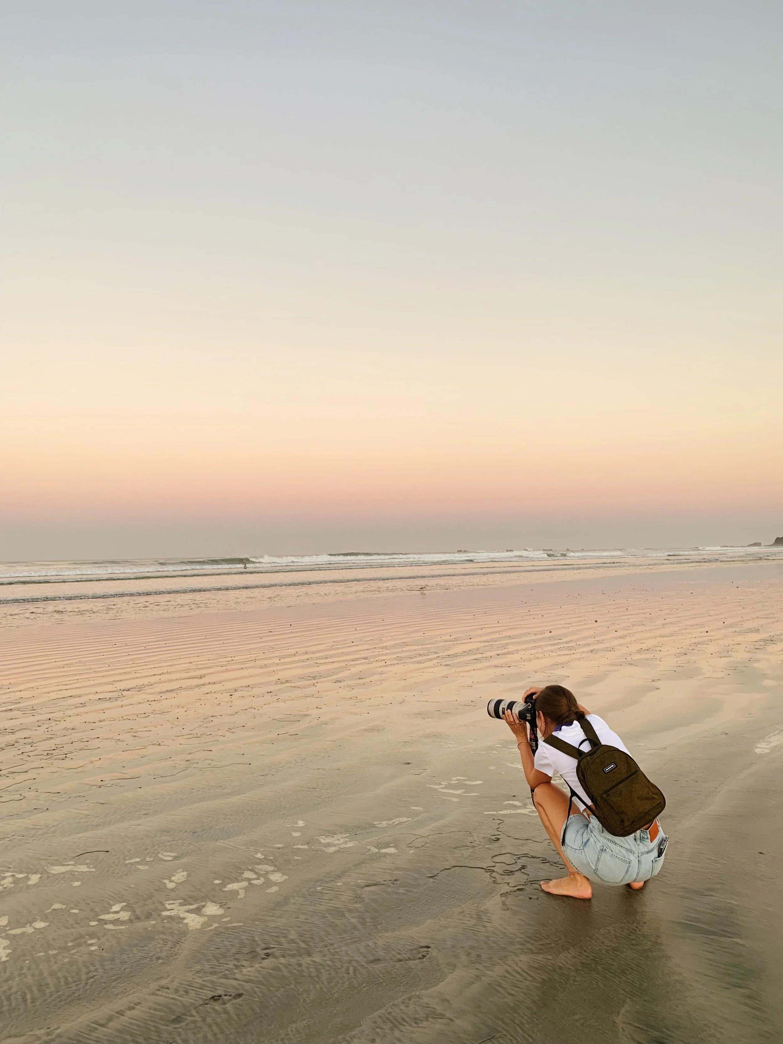 Woman crouching on the beach, taking photos with a camera during sunset, wearing a white shirt, denim shorts, and a backpack.