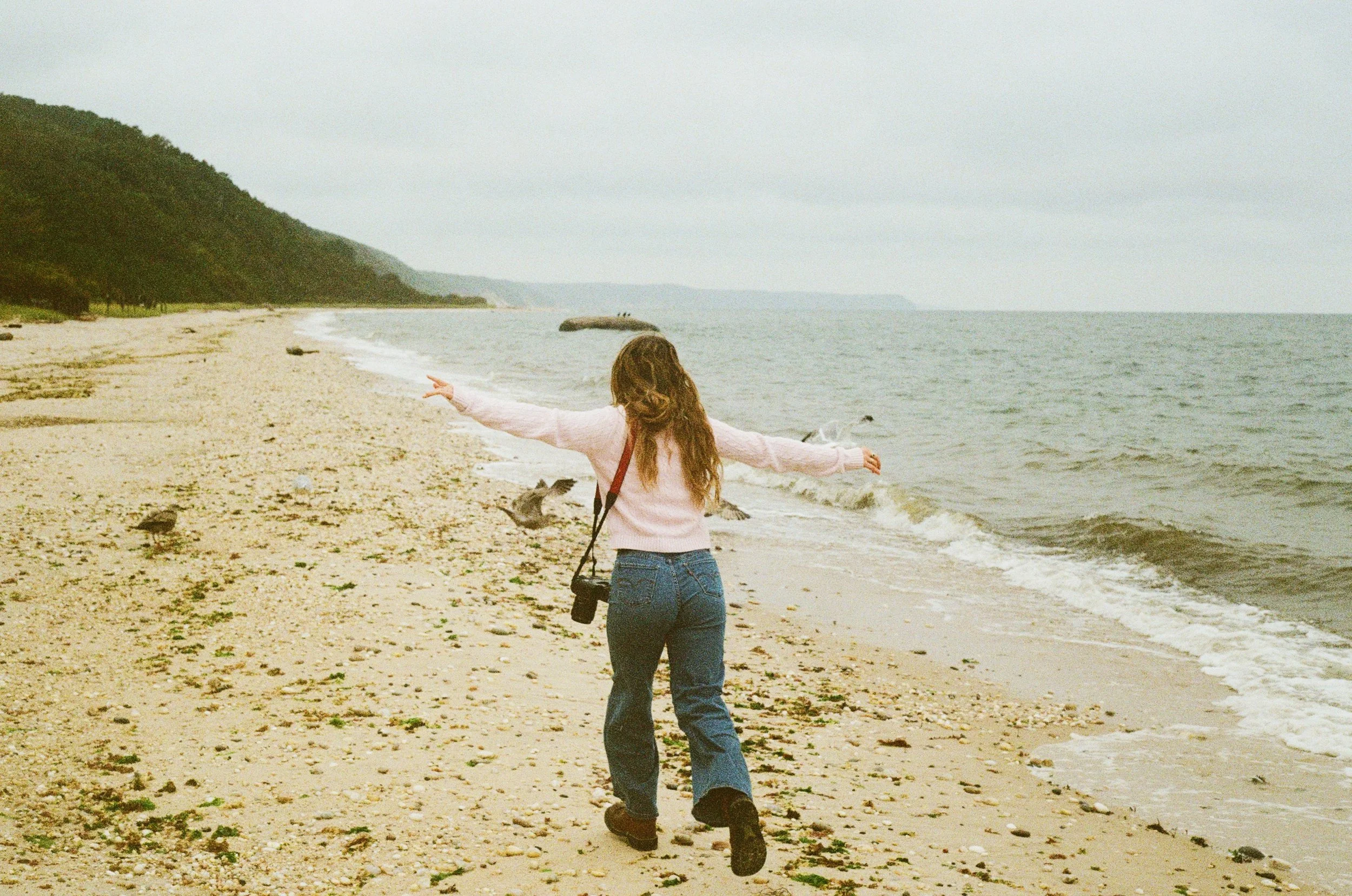 A woman walking along a sandy beach with her arms outstretched, birds flying nearby, overcast sky, distant cliffs, and calm ocean waves.
