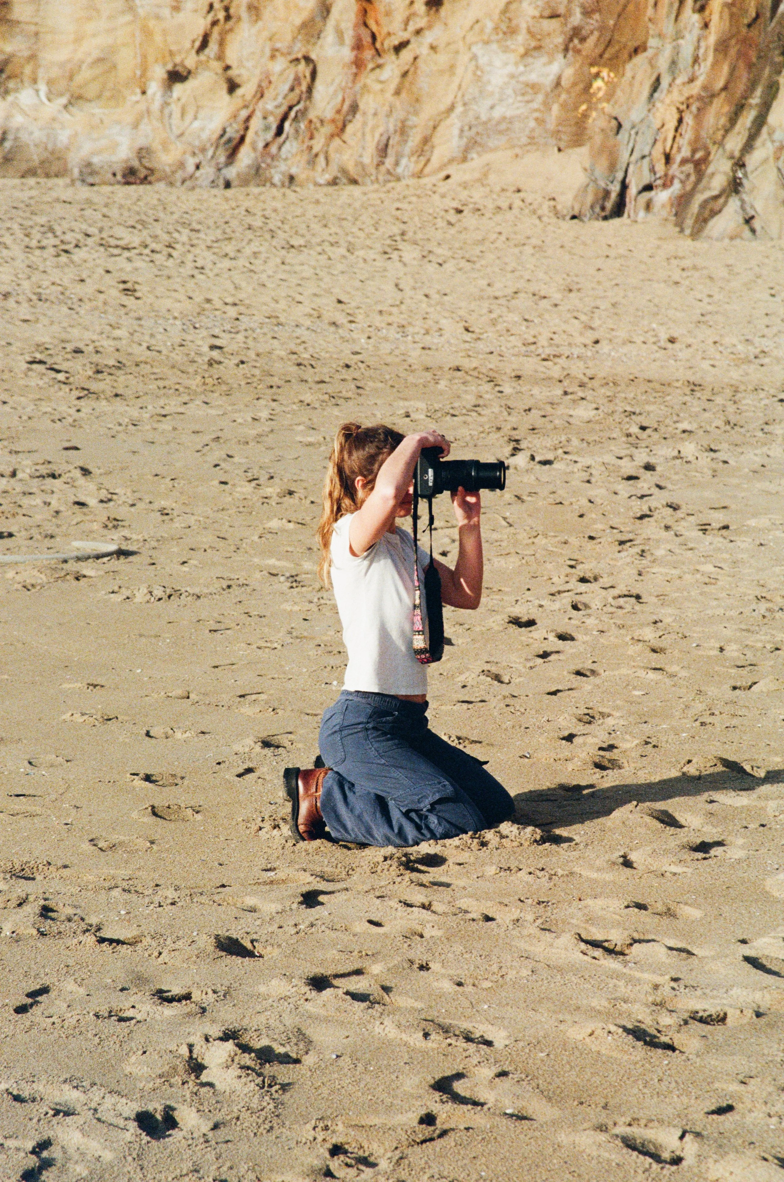 A young girl kneeling on a sandy beach, looking through a camera or binoculars, with rocky cliffs in the background.