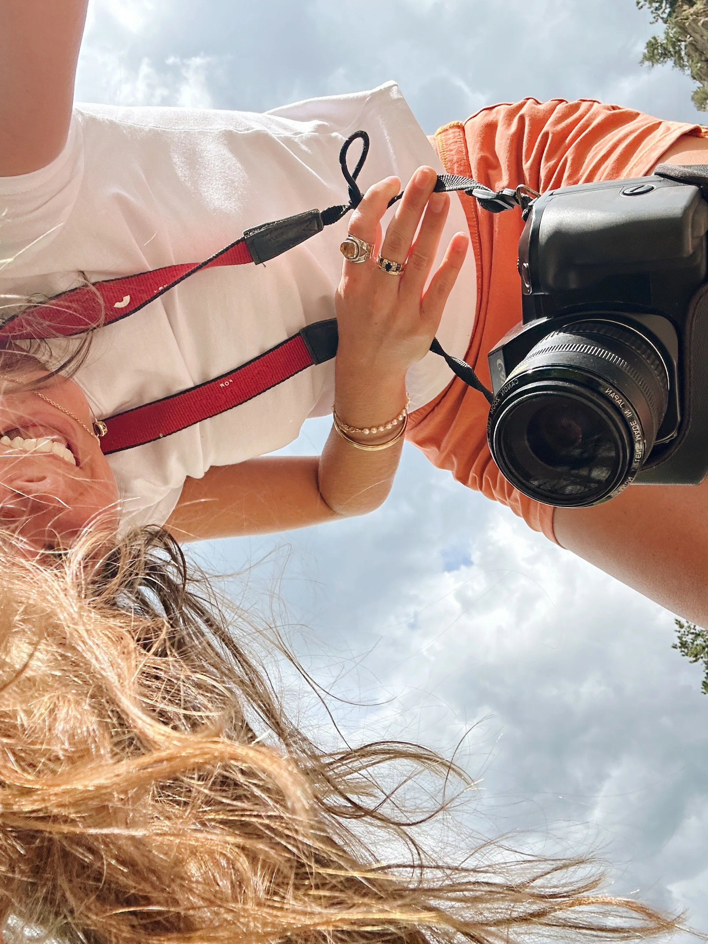 Photograph of a woman with curly blonde hair taking a selfie outdoors with a camera, smiling, sky and clouds in the background.