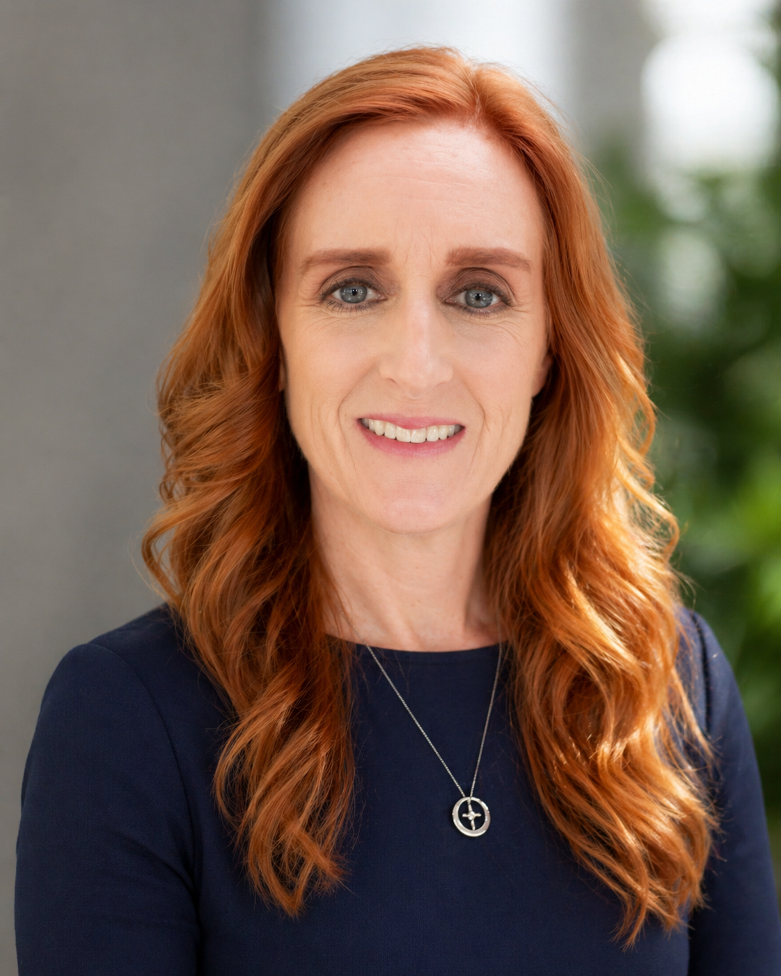A woman with long wavy red hair and blue eyes, wearing a dark blue top and a silver necklace with a cross pendant, smiling indoors with a blurred background of green plants.