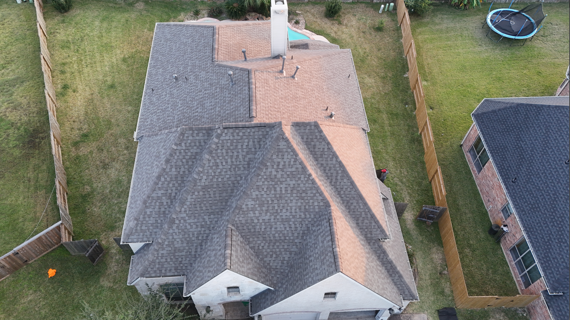 Aerial view of a house with a multi-colored roof, surrounding fenced yard, trampoline, and a person near the house.