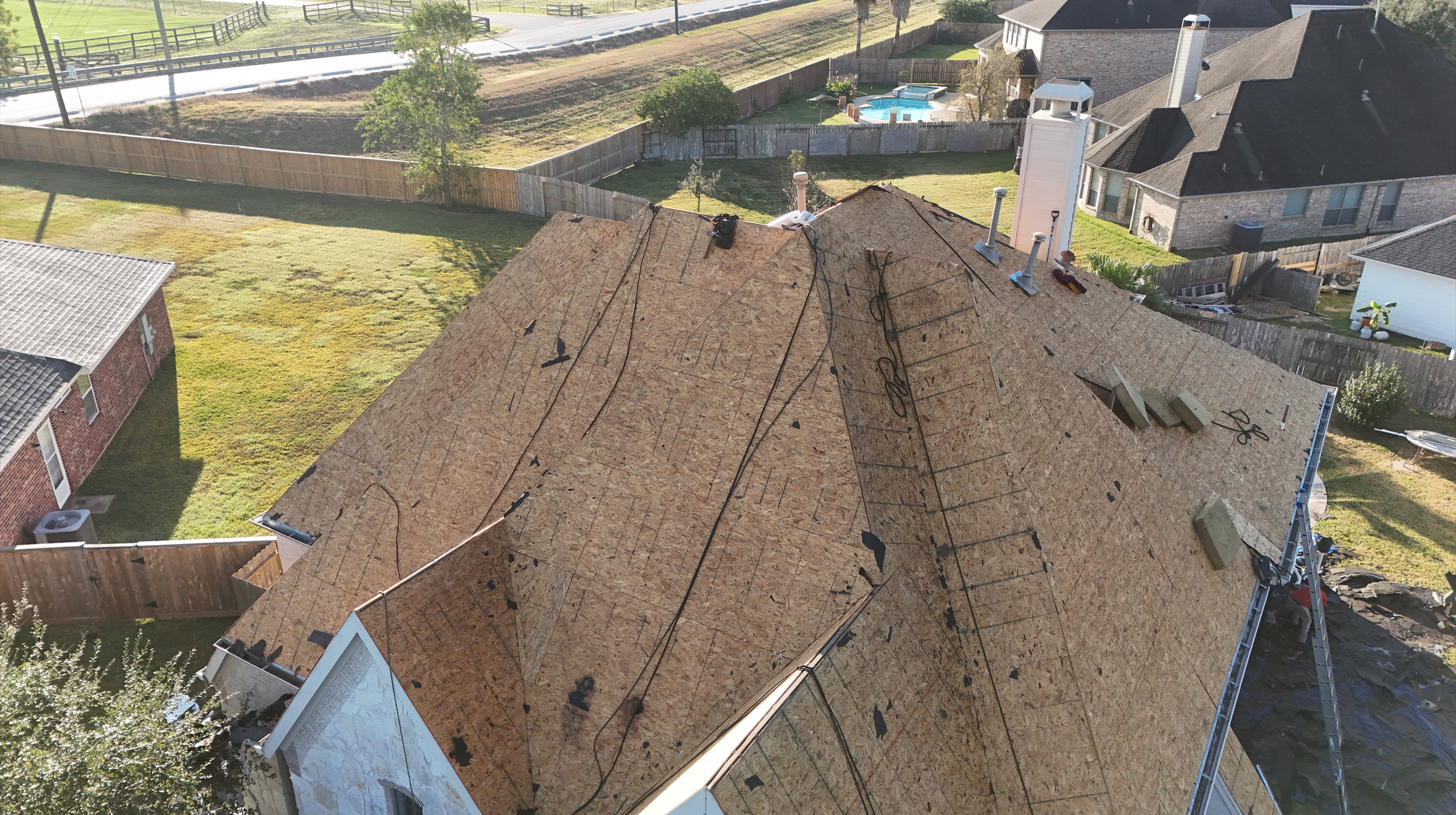 Aerial view of a house undergoing roof replacement with plywood sheathing installed, with electrical wires and construction tools visible, surrounded by fenced yards and neighboring houses.