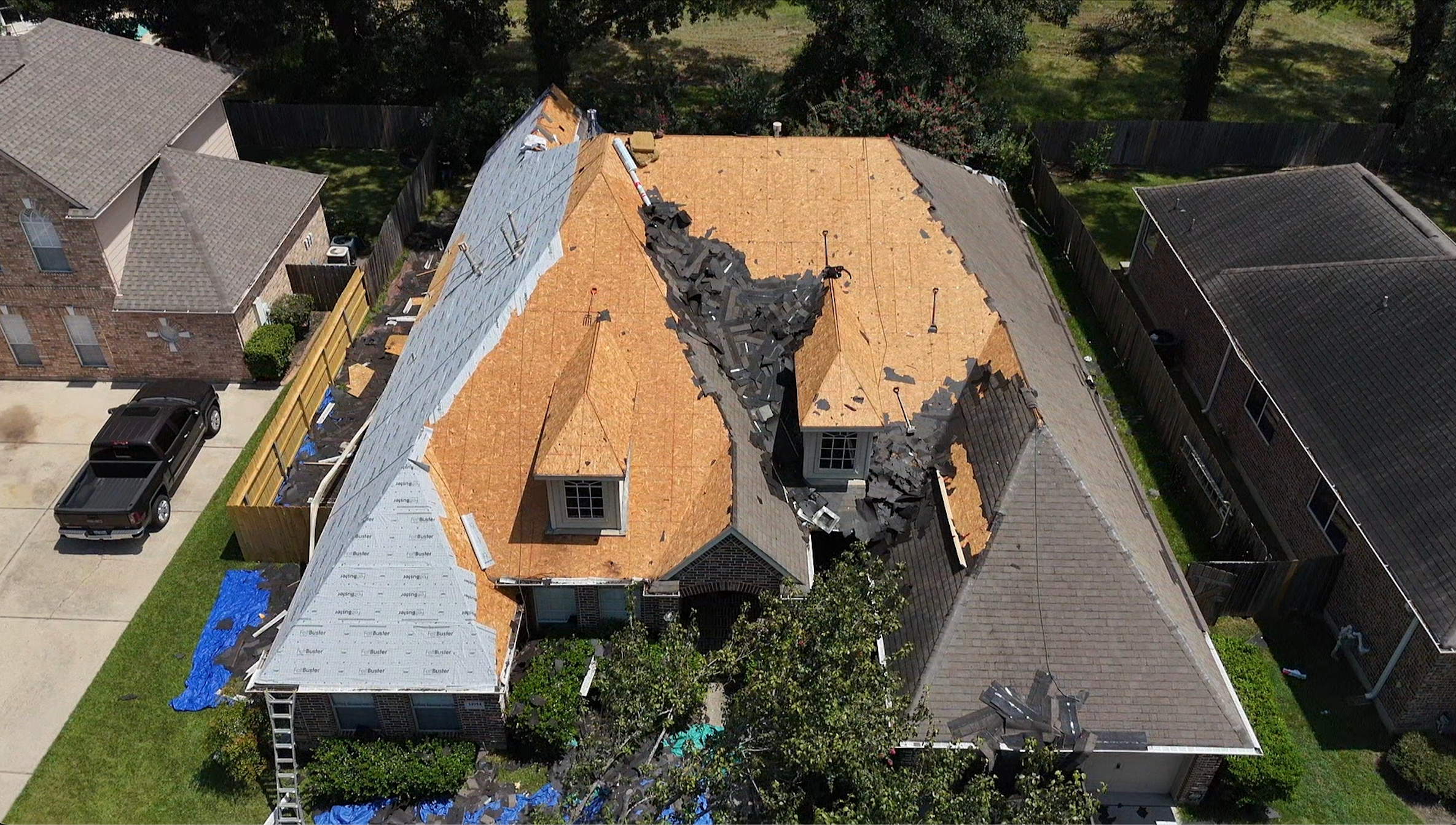 A house undergoing roof repair or replacement with part of the roof torn off, exposing the interior and roofing materials scattered across the roof and yard, neighboring houses nearby.