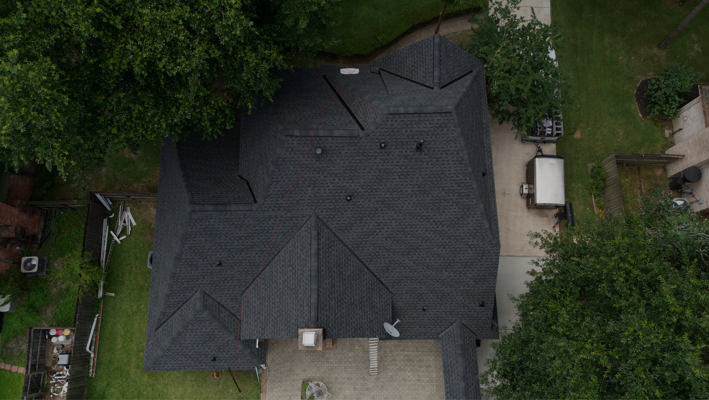 Aerial view of a house with dark gray roof, surrounded by green trees and a back yard with a paved area, a small storage shed, and outdoor furniture.
