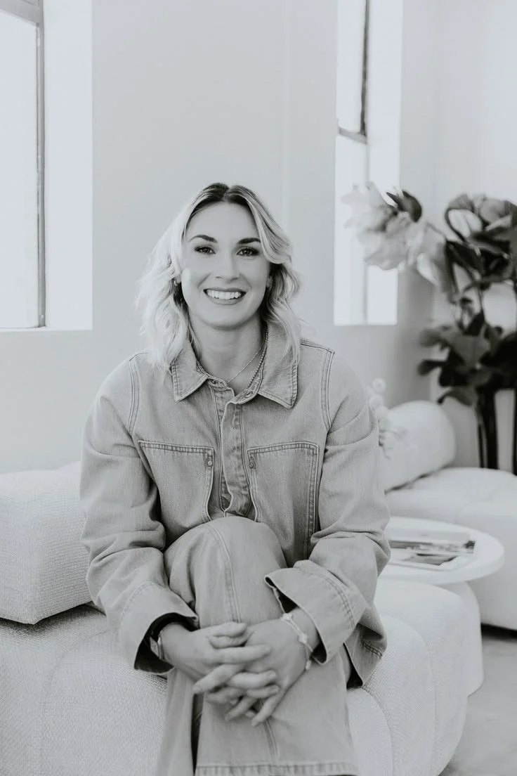 A young woman with blonde hair sitting on a sofa in a bright room, smiling at the camera.