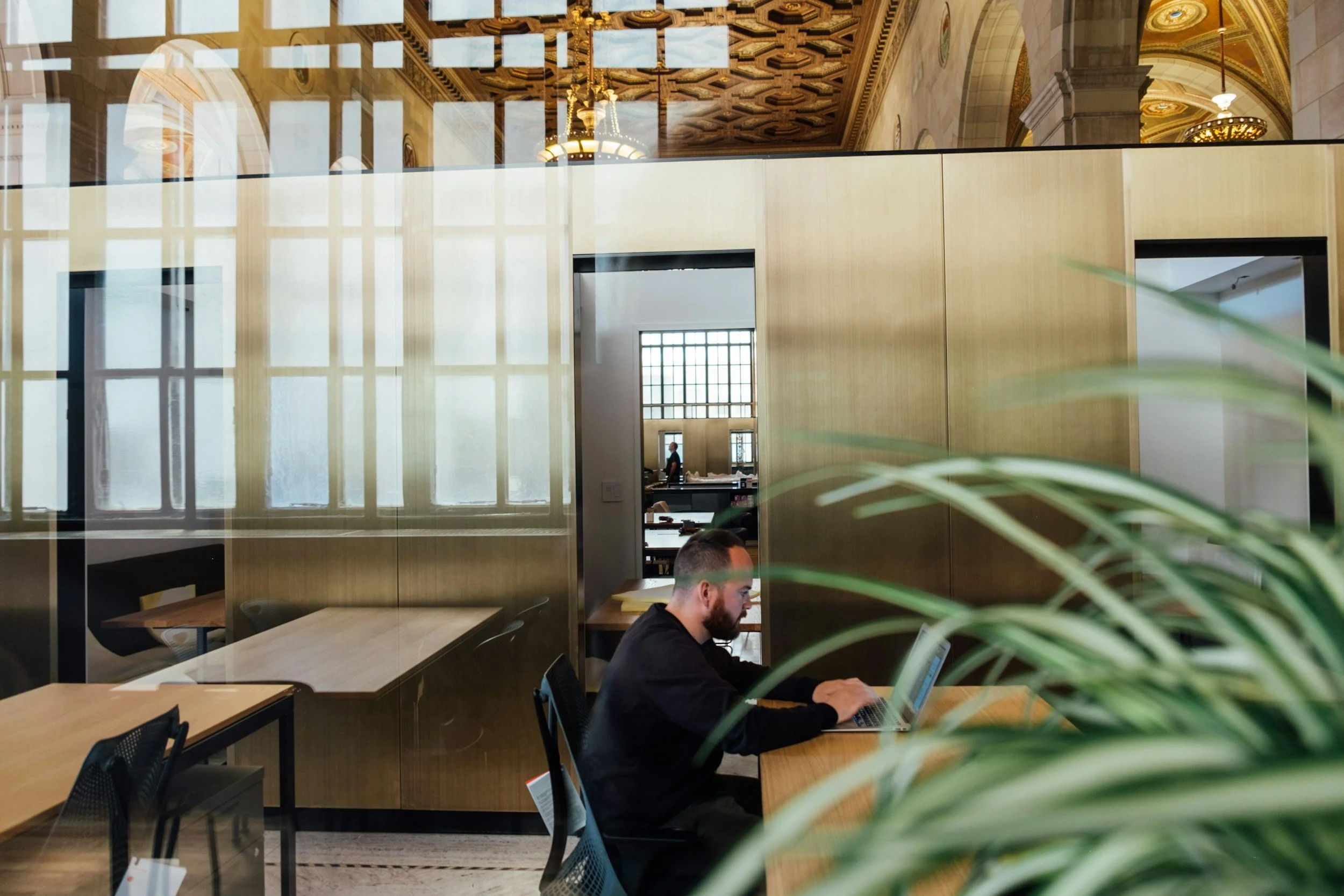 En mand arbejder på laptop i et moderne kontormiljø med store vinduer og historisk arkitektur i baggrunden.