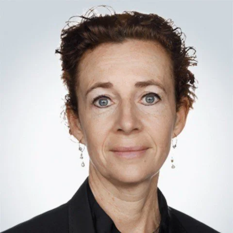 Portræt af en kvinde med kort, krøllet brunt hår og blå øjne iført sorte øreringe og sort tøj.