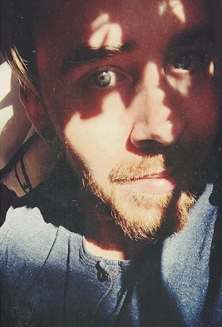 Close-up of a young man with light-colored eyes, light facial hair, and short hair, partially illuminated by sunlight with shadows on his face.