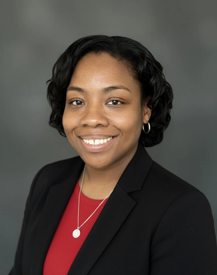 Professional portrait of a smiling Black woman with short curly hair, wearing a black blazer, red top, hoop earrings, and a silver necklace with a pendant, against a gray background.