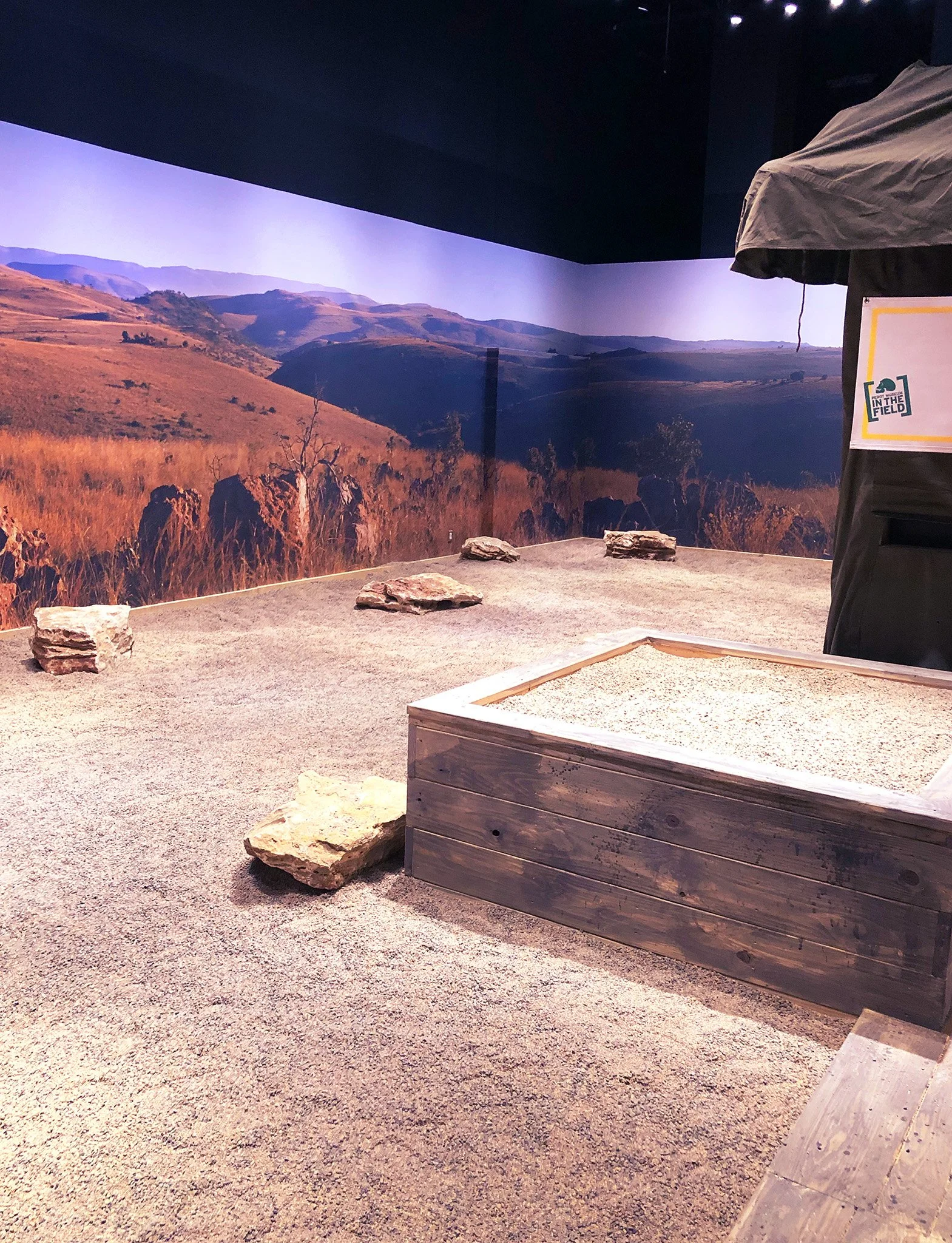 Indoor exhibit with a large mural of rolling hills and mountains in a rural landscape, a dirt floor with large rocks, and a wooden sandbox in the foreground.