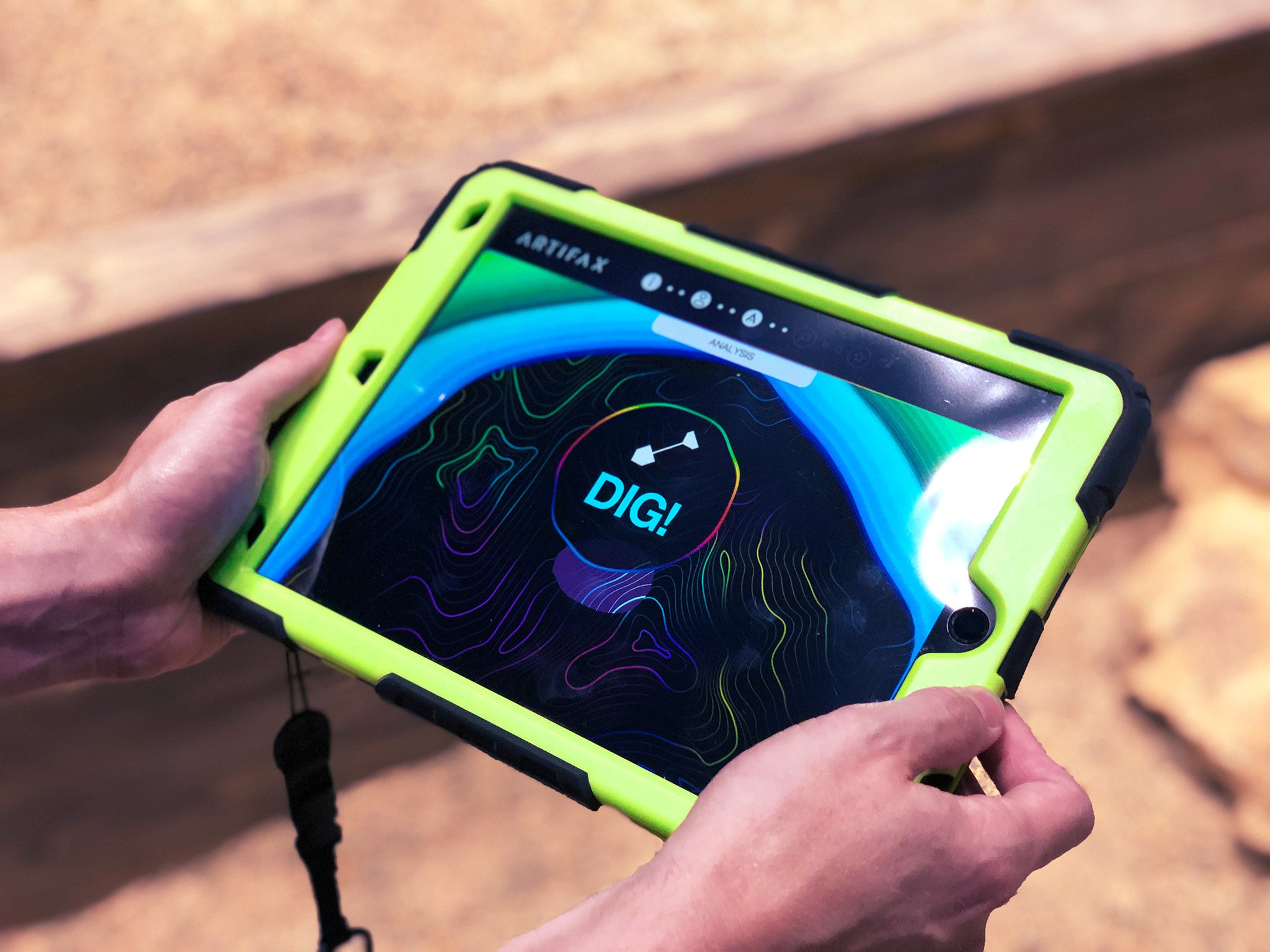Close-up of a person holding a yellow and black electronic device with a colorful, topographical map and the words 'DIG!' displayed on the screen.