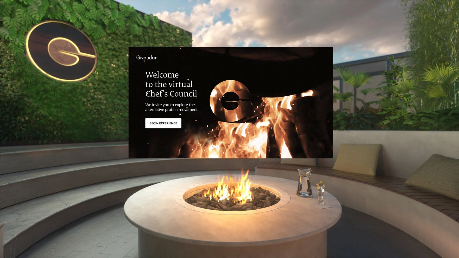 Outdoor patio with a fire pit table, surrounded by curved built-in seating with cushions, and glass pitchers on the table. Green plants and trees in the background, with a partial view of a modern building and a circular logo on the wall. Large digit
