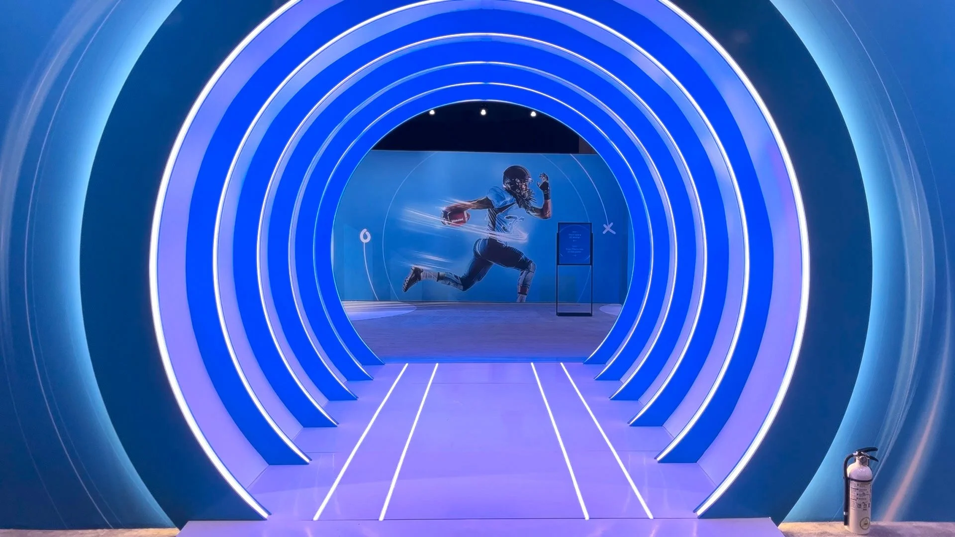 Blue illuminated tunnel leading to a display area with a large mural of a football player running, holding a football, in a modern exhibit or museum.