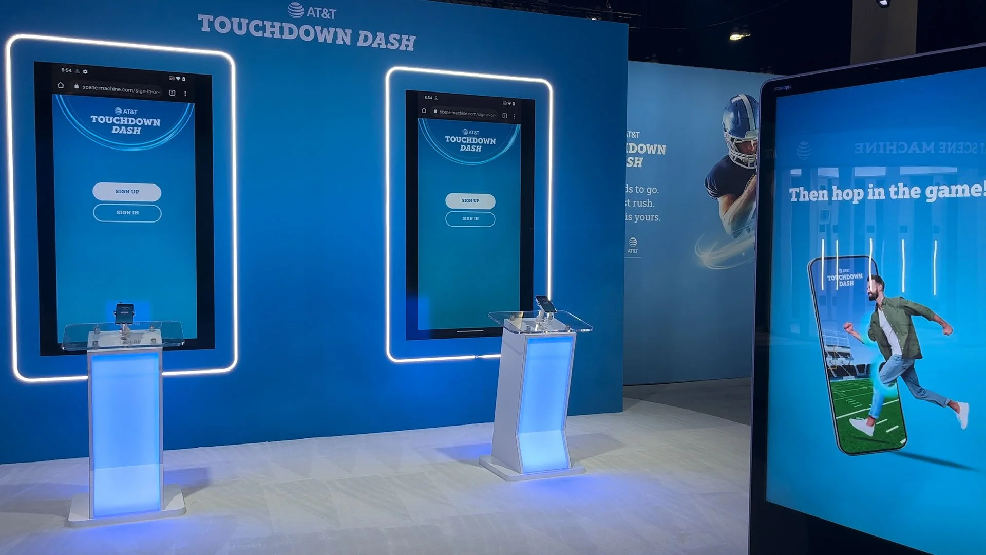 Display booth for AT&T's Touchdown Dash, featuring two digital screens with app sign-up options, a football-themed backdrop, and a large poster of a man running with a smartphone in a football stadium.