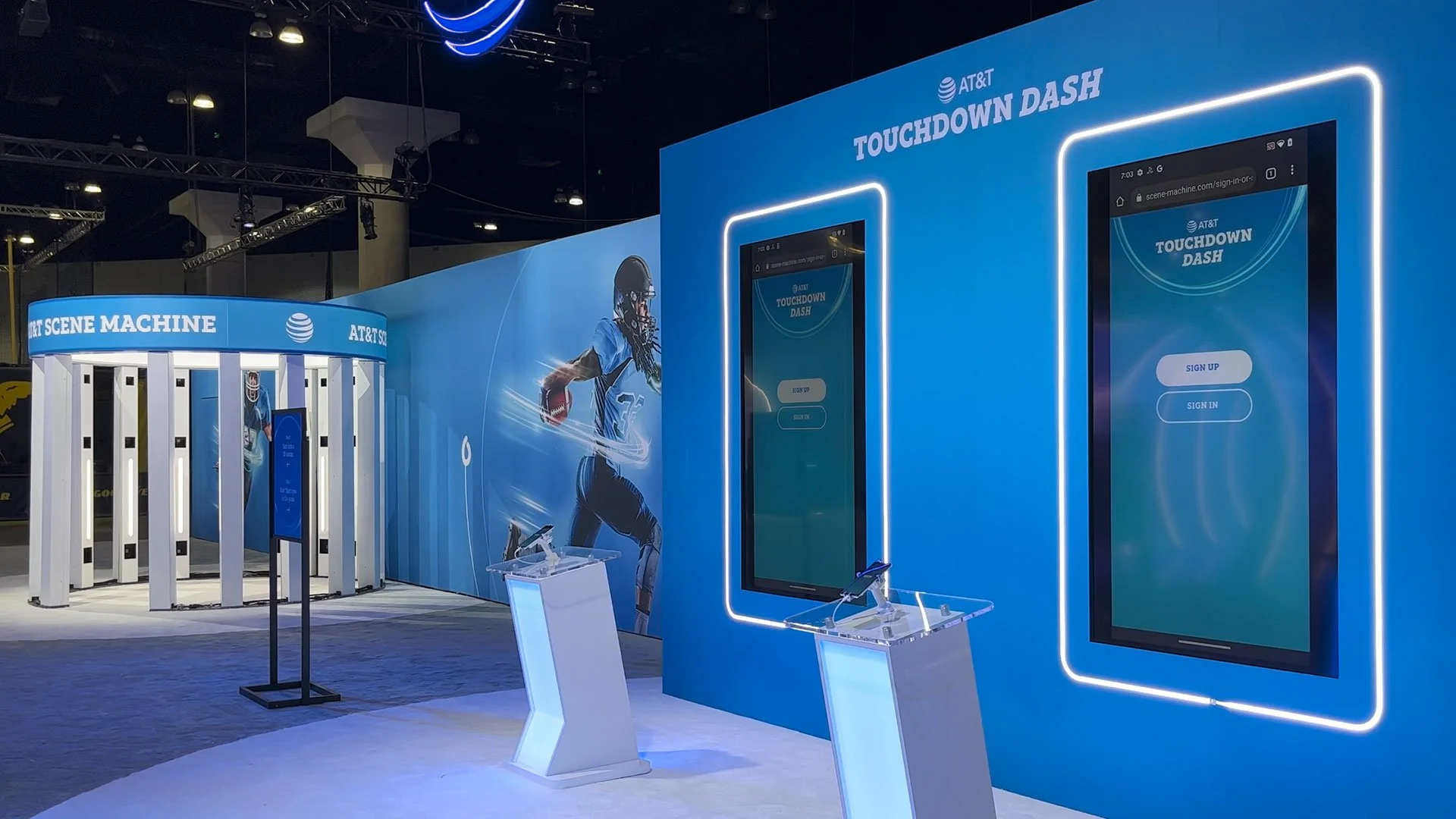 AT&T booth featuring a large blue wall with a digital sign-in screen for the 'Touchdown Dash' game, a football player illustration, and a curved white structure with game activity stations, all illuminated with bright lighting.