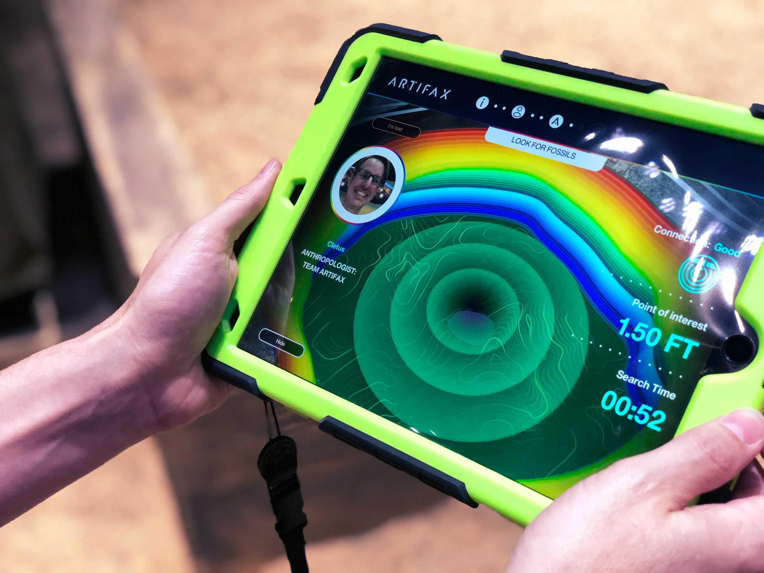 Close-up of a person holding a tablet device with a green protective case, displaying a colorful radar or topographical map on the screen, with some text and a small profile picture of a man.