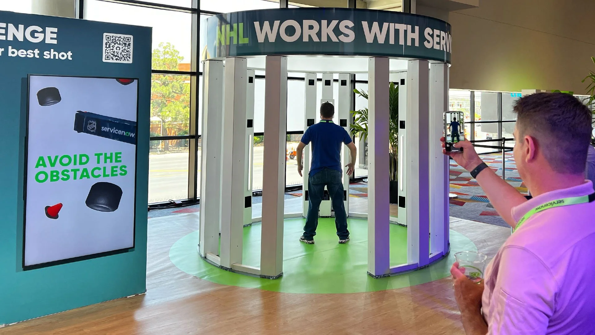 Two men at an indoor event attempting to pass through a circular obstacle course, with one man using a smartphone to take a photo or video.