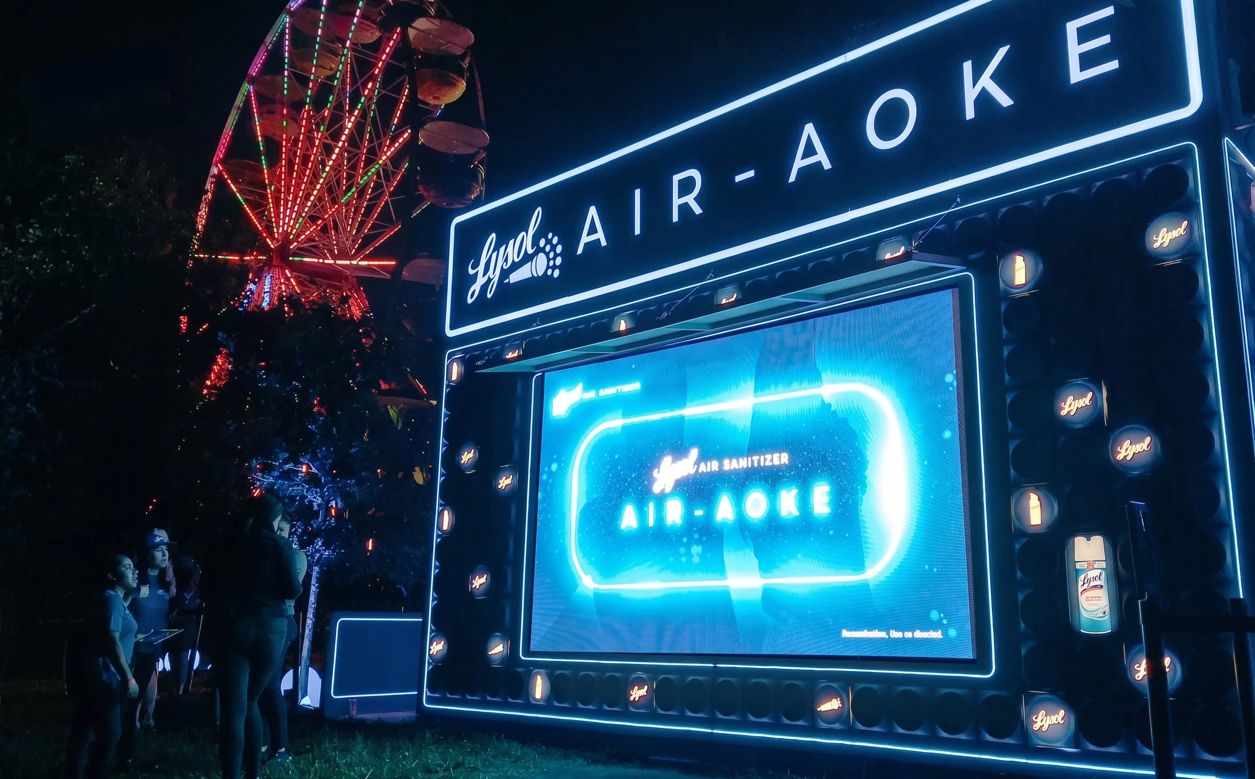 People standing in front of a large illuminated sign with the Lyod air-aoke advertisement at night, and a Ferris wheel in the background.