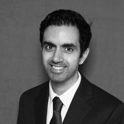 Headshot of a young man with dark hair, wearing a dark suit, white shirt, and green tie, smiling against a gray background.