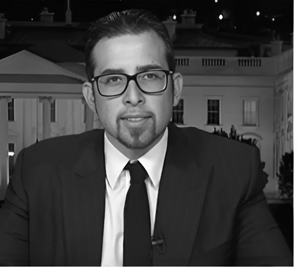 A man wearing glasses and a black suit with a white shirt and black tie sitting in front of a background resembling the White House at night.